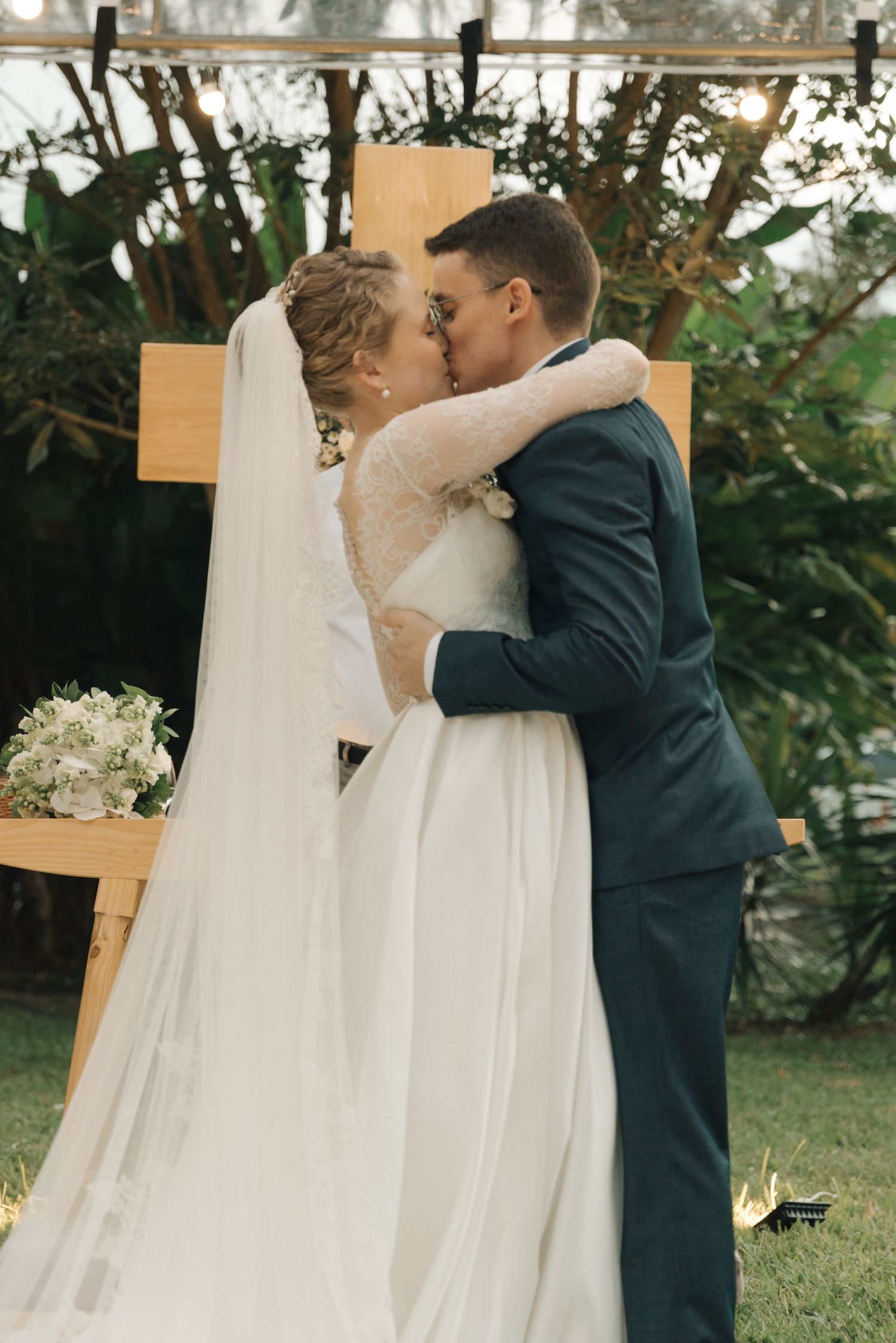 Intimate wedding ceremony moment with wooden cross backdrop and string lights in garden setting.