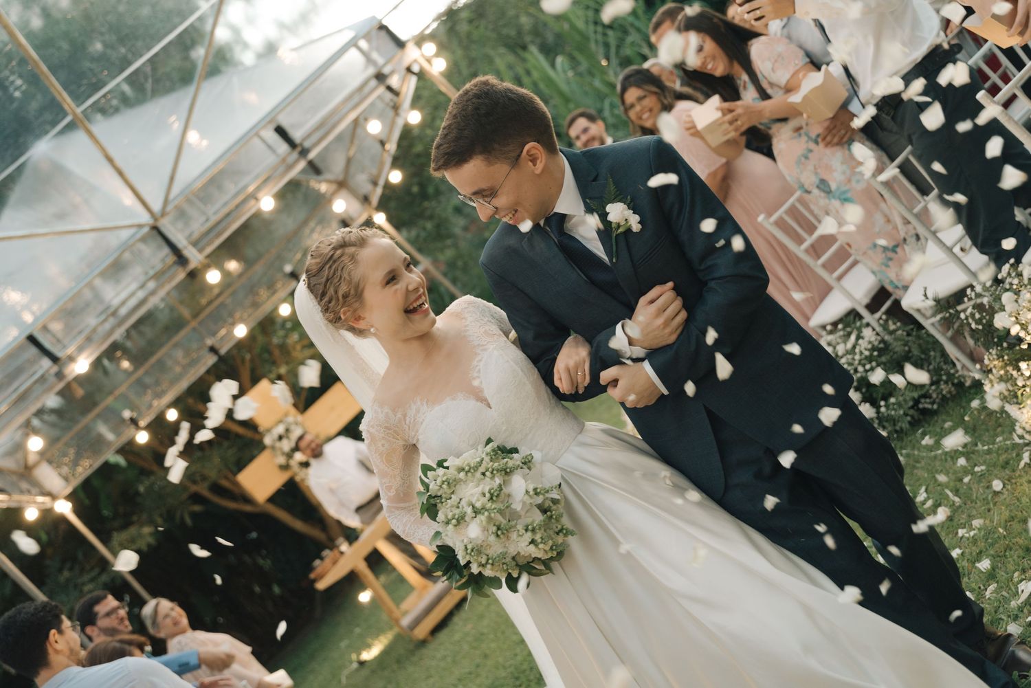 A joyful wedding couple walks through shower of white petals under a clear tent with string lights.
