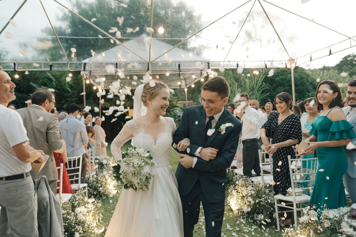 Wedding couple walks through floral-lined aisle as guests celebrate under clear tent reception venue.