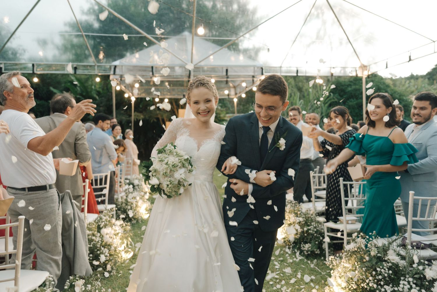 Newlyweds walk down aisle under clear tent while guests throw petals at outdoor wedding reception.