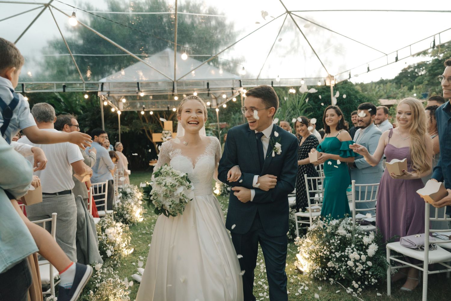 Happy couple proceeds down decorated aisle while wedding guests throw petals during outdoor celebration.