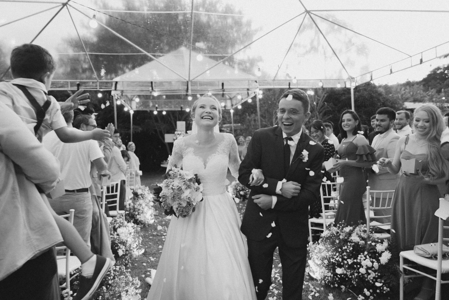 Black and white photo of newlyweds walking down aisle during outdoor tented wedding ceremony.