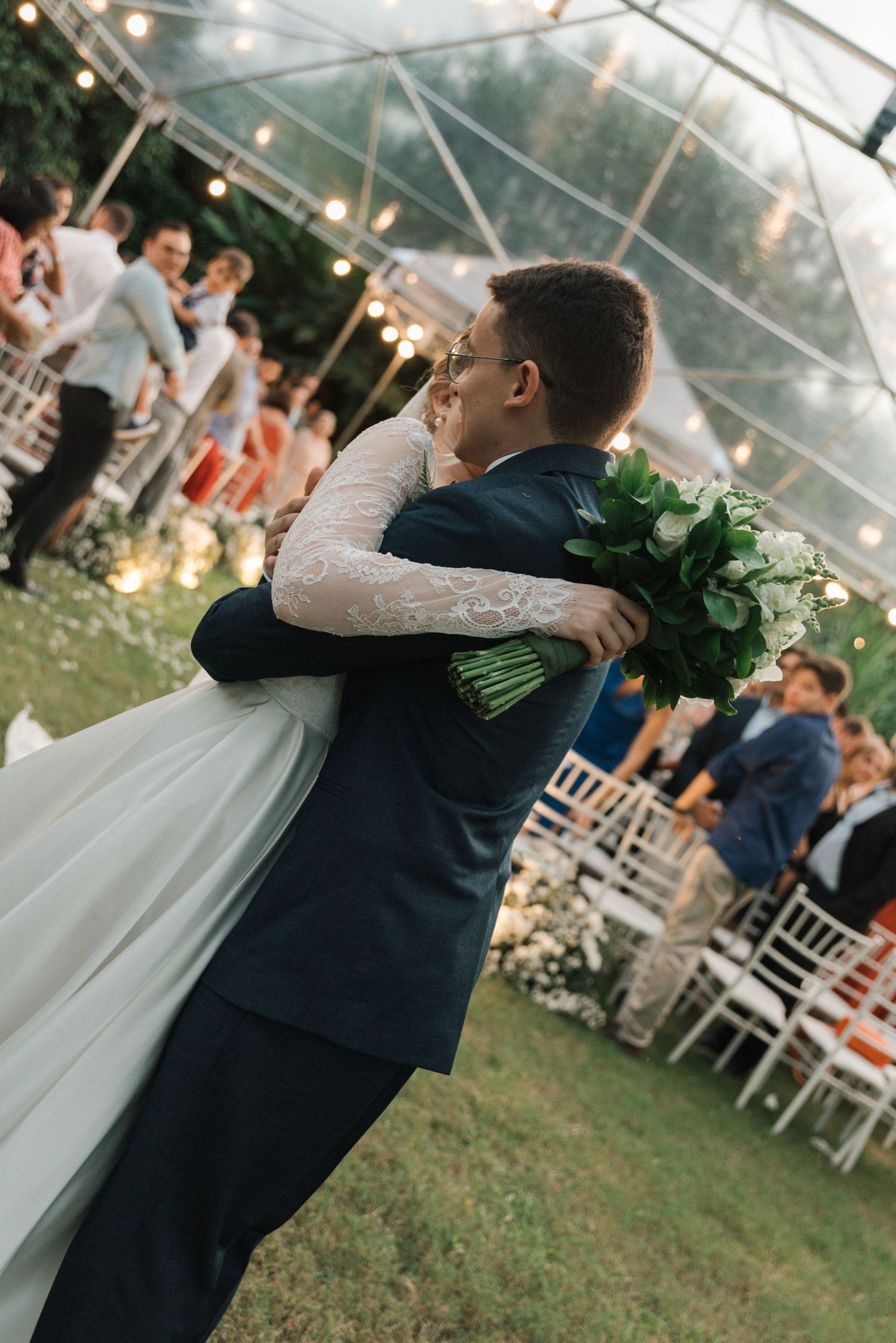 Bride and groom share intimate embrace while holding bouquet during outdoor reception.