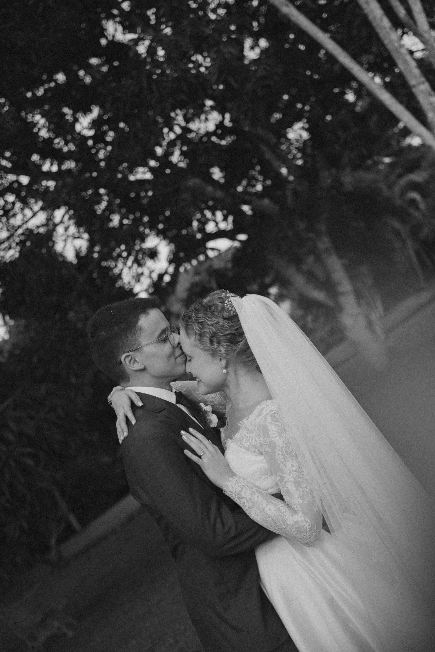 Black and white photo of romantic wedding couple embracing under tree branches.