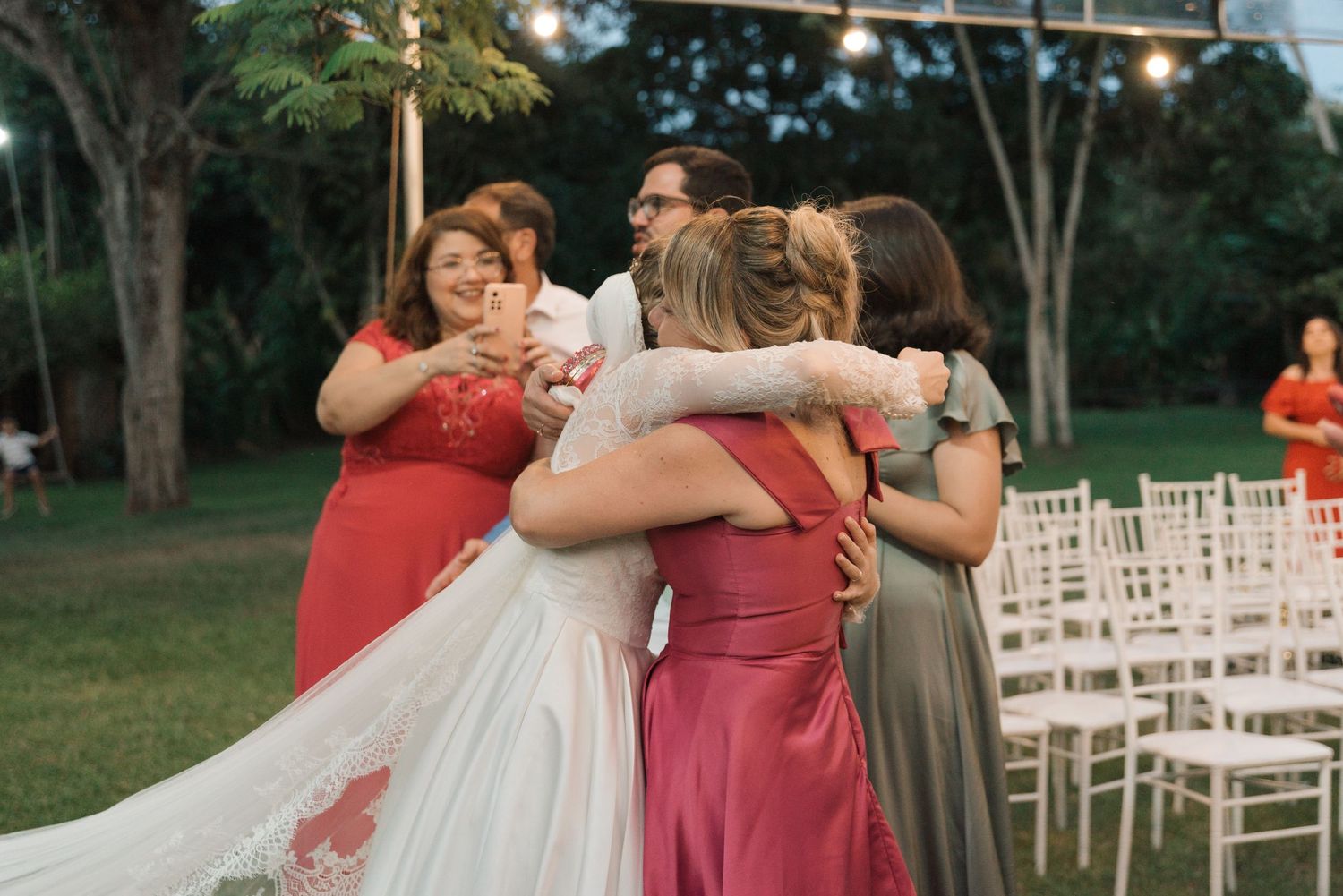 Bride in white dress receives group hug from wedding party in coral dresses.