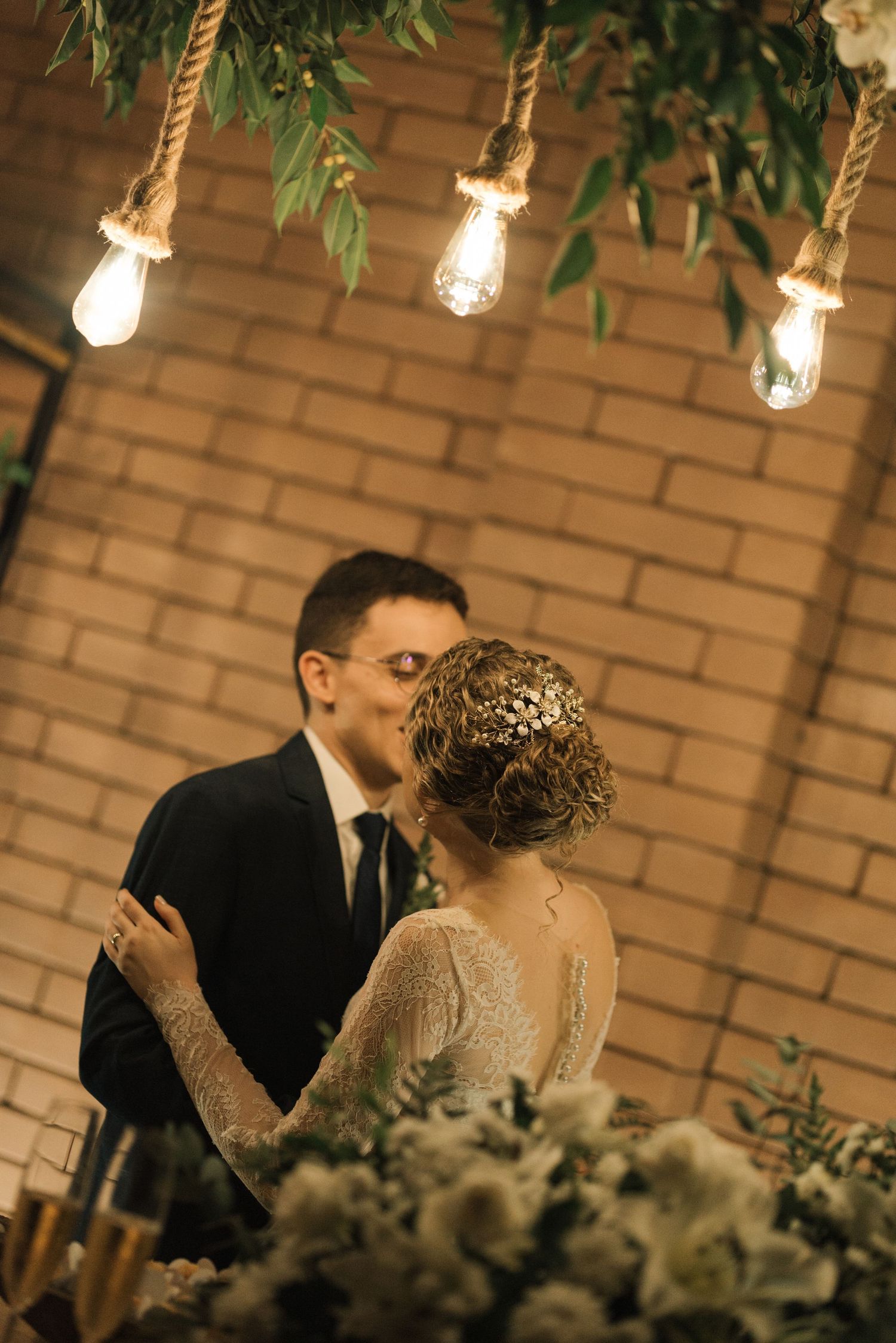 Romantic wedding scene with hanging Edison bulbs and greenery against brick backdrop.
