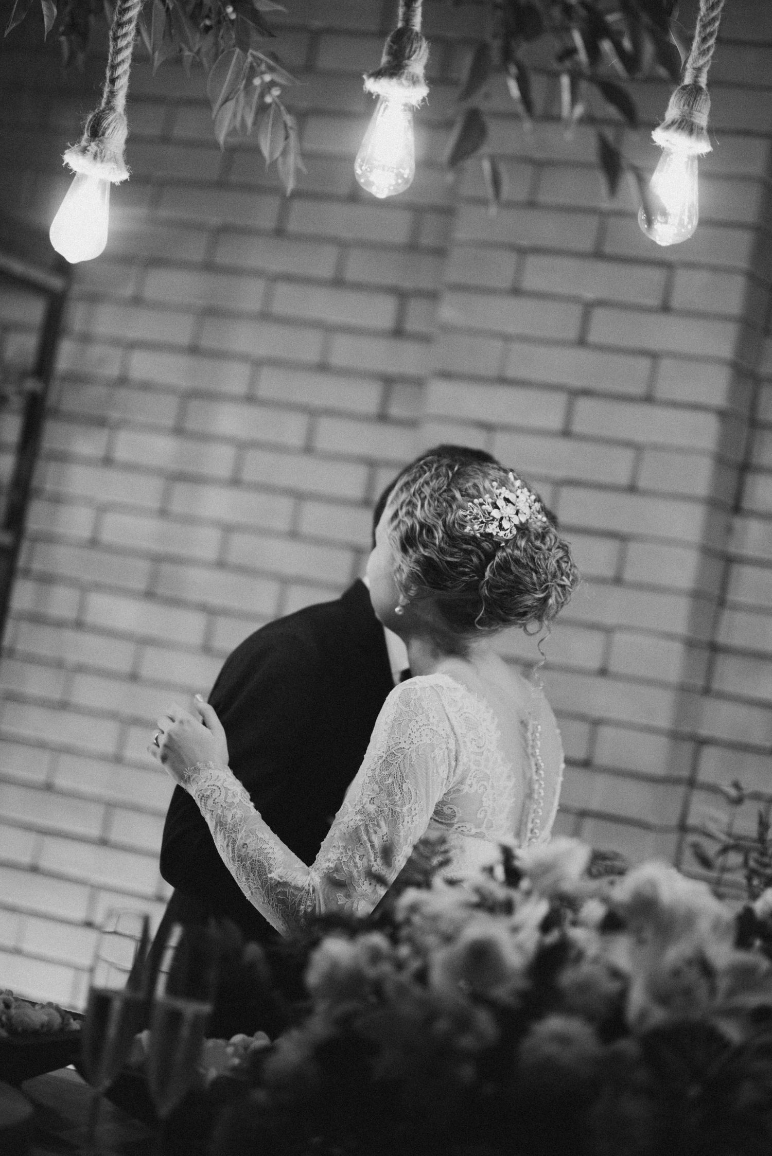 Black and white photo of newlyweds dancing beneath vintage string lights.