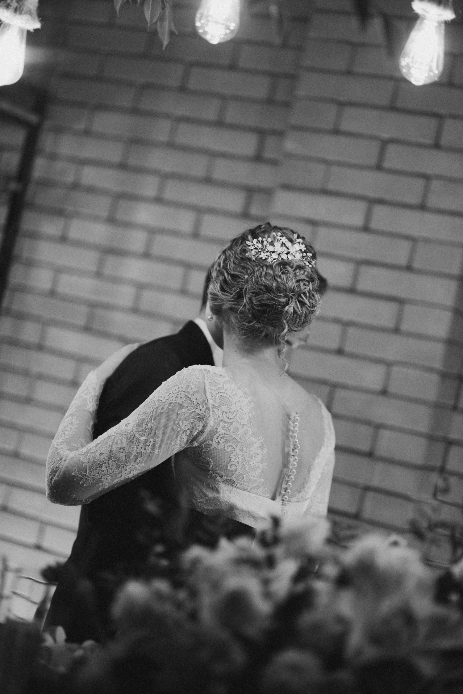 Back view of bride in lace dress dancing with groom under suspended lights.