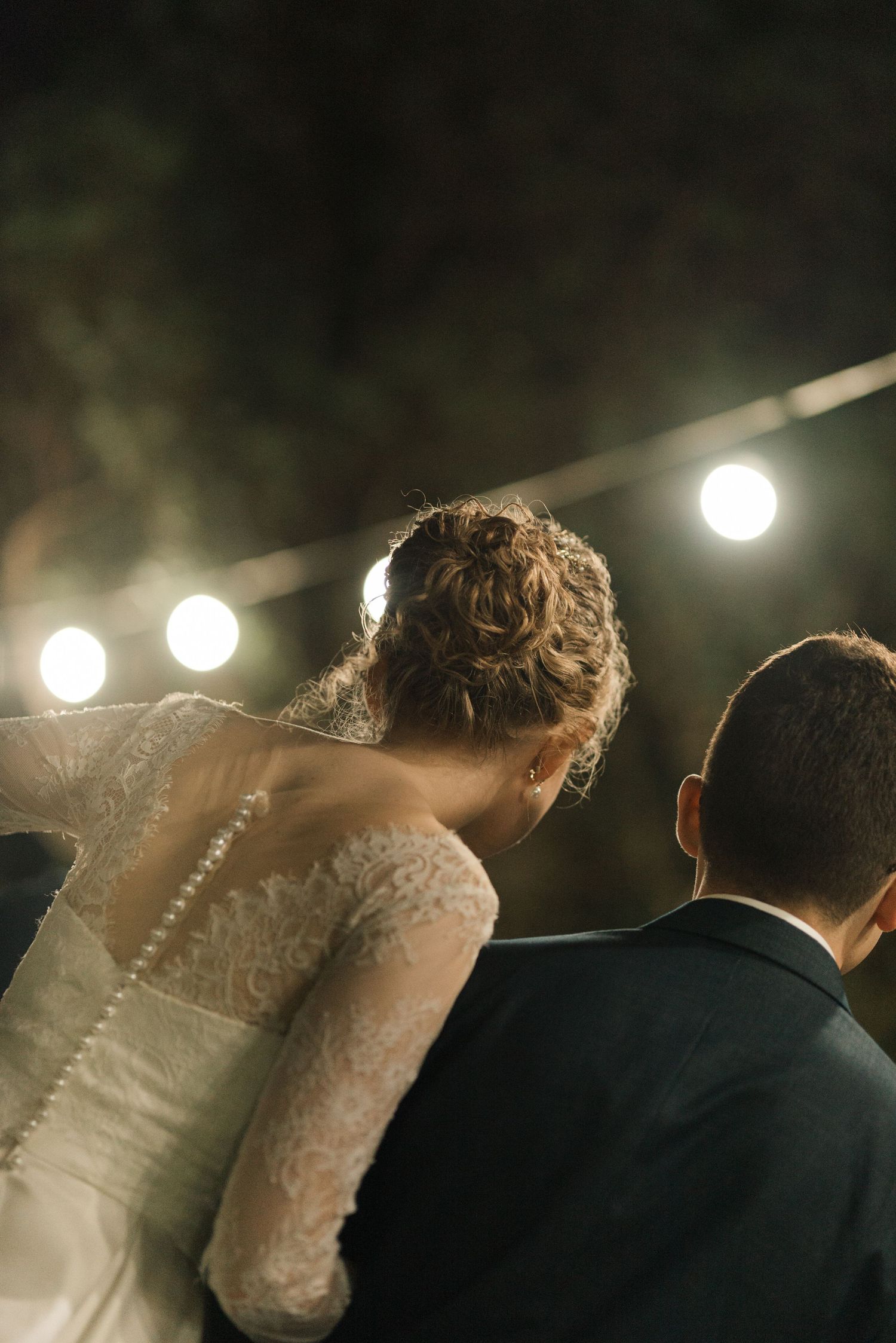 A wedding couple's back view showing intricate lace details of the bride's dress under overhead lights.