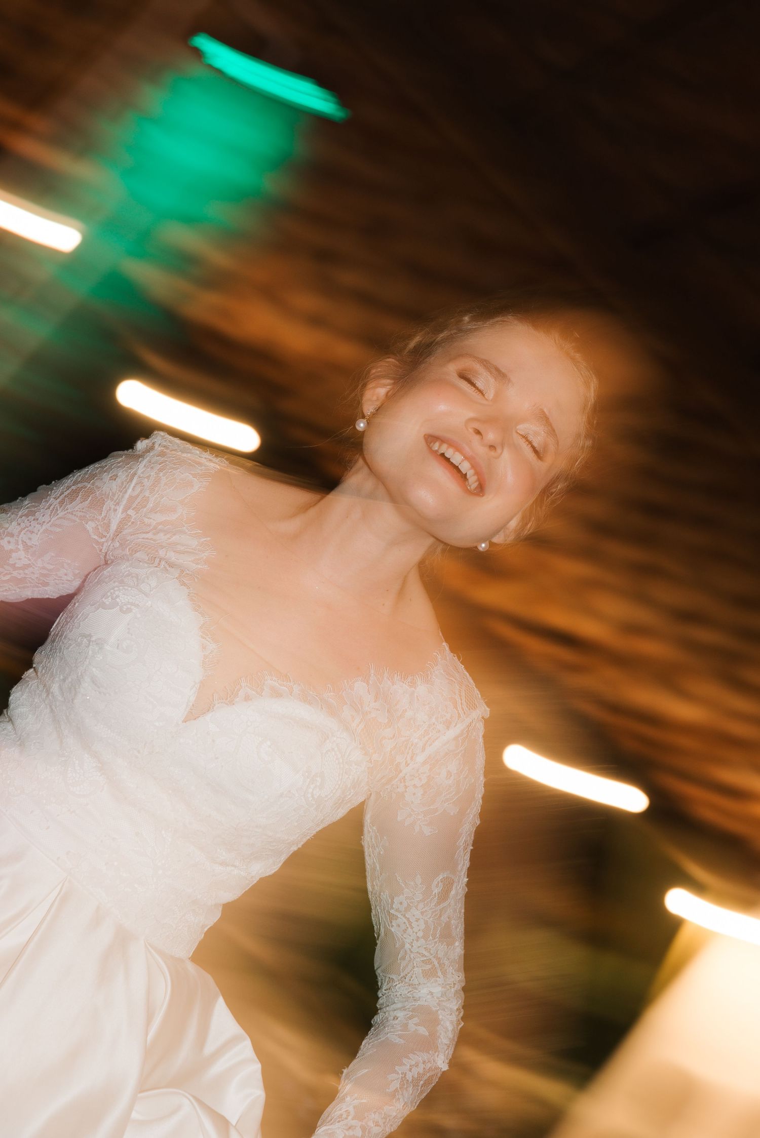 A bride in a white lace dress dances joyfully under green and warm lighting at a wedding reception.
