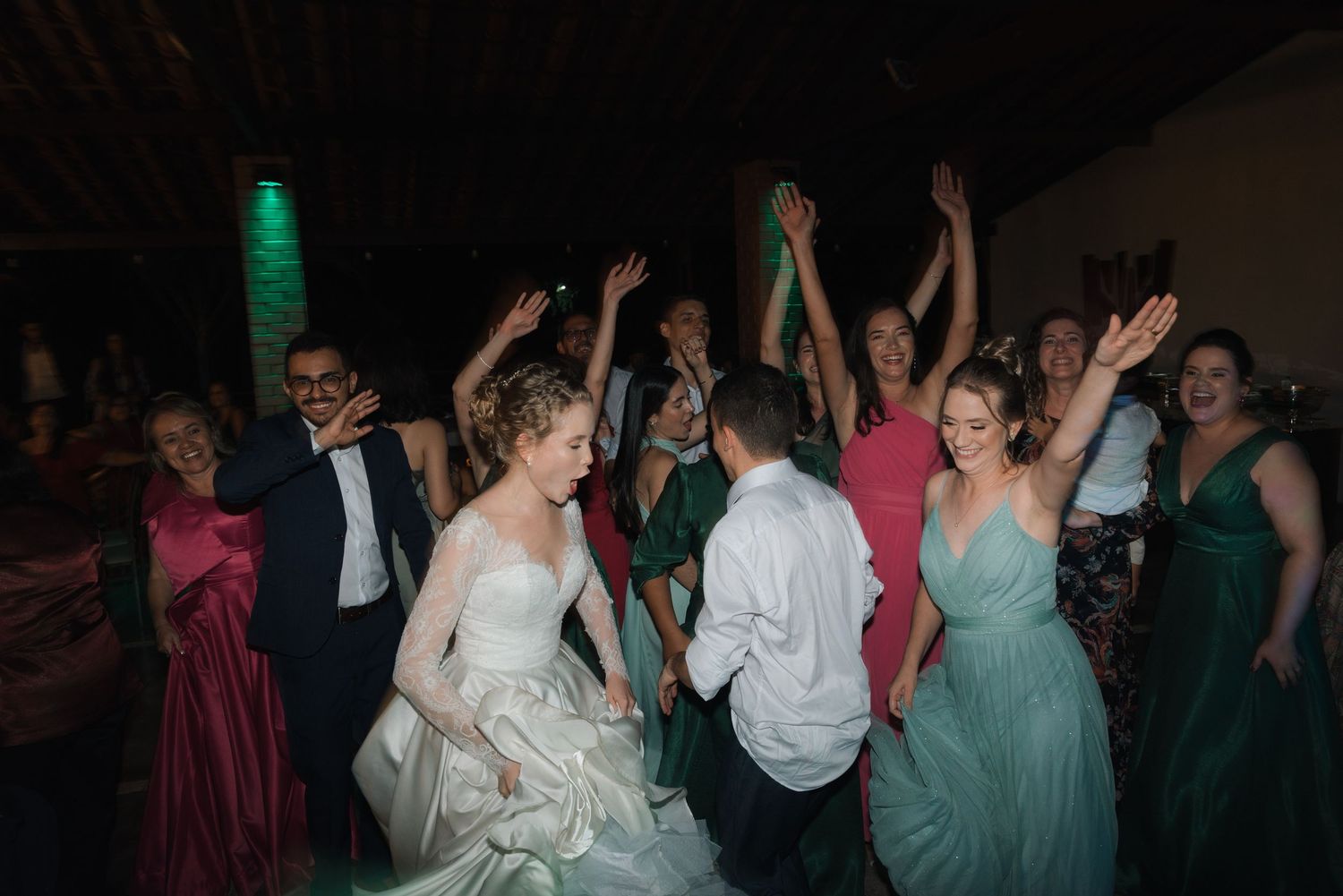 Wedding guests dancing with raised arms under green accent lighting at reception venue.