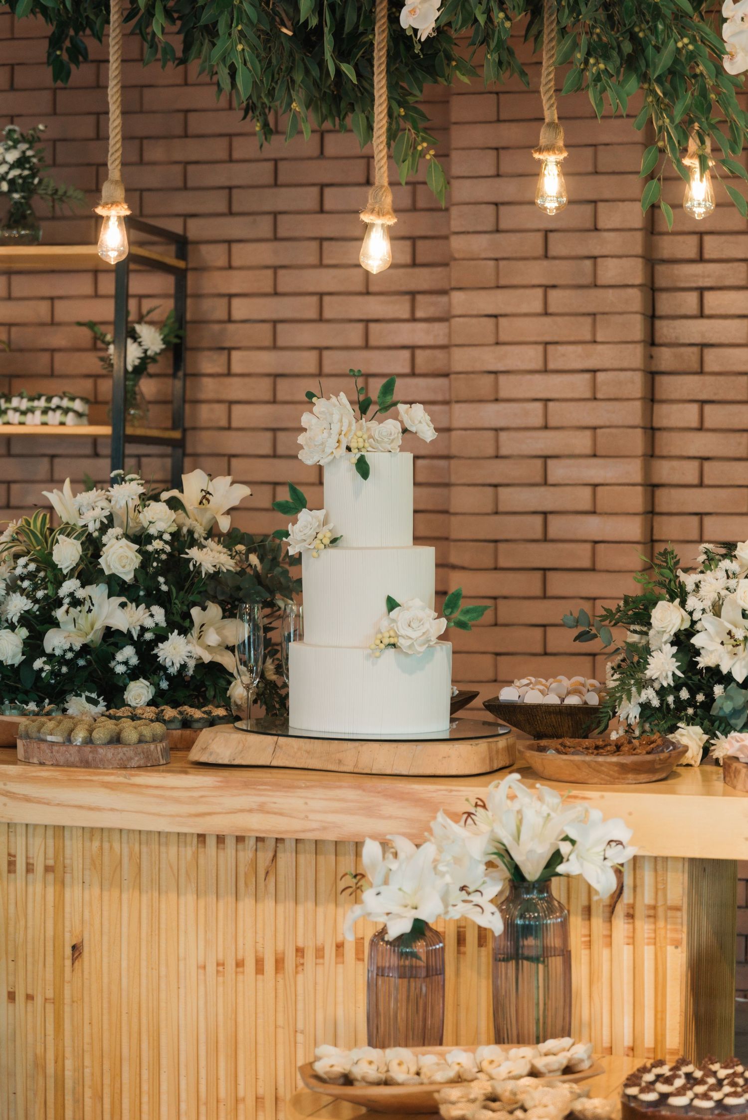 Elegant white wedding cake display with hanging lights and floral decorations against brick wall.