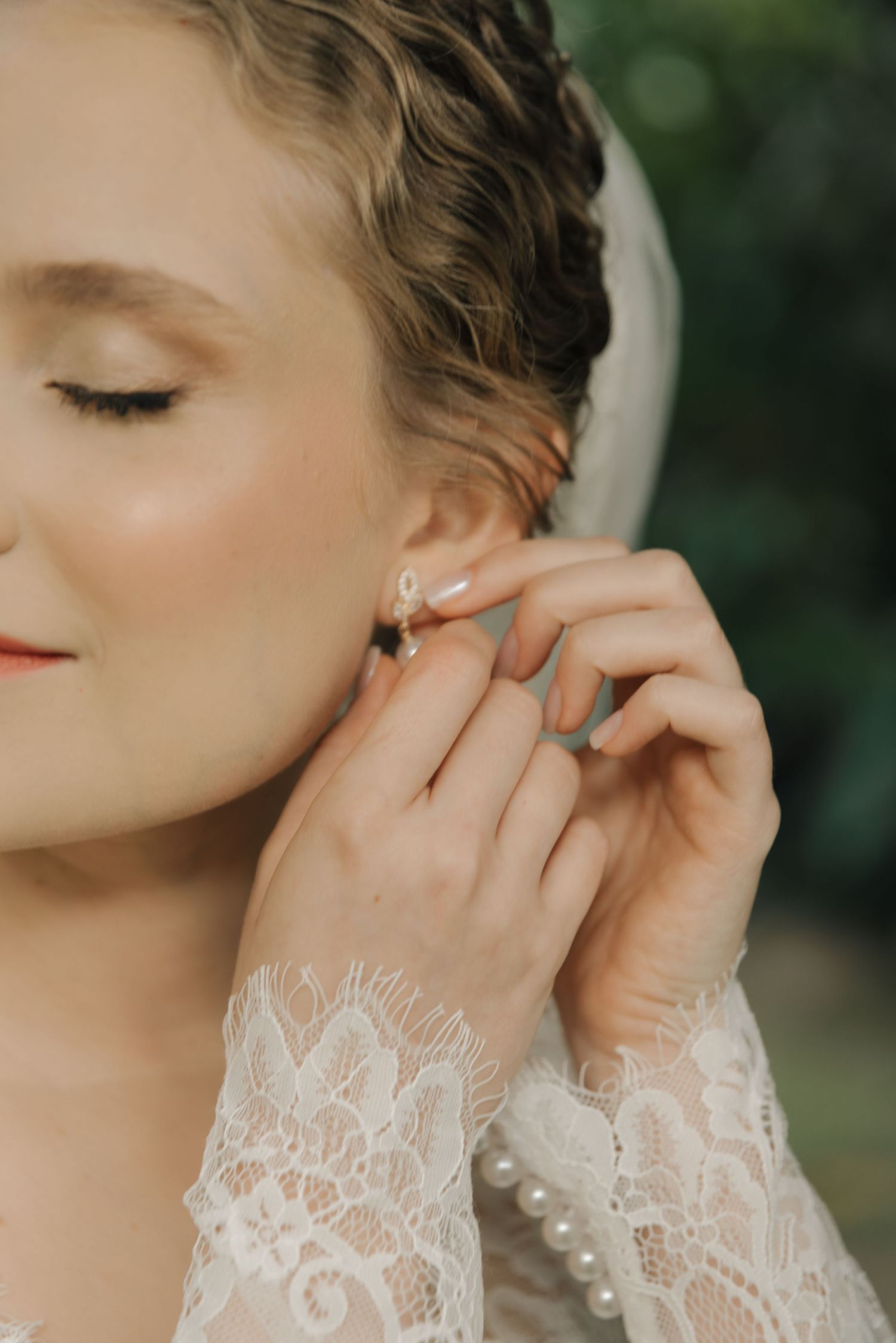 Close-up of bride adjusting pearl earring while wearing lace sleeve wedding dress.