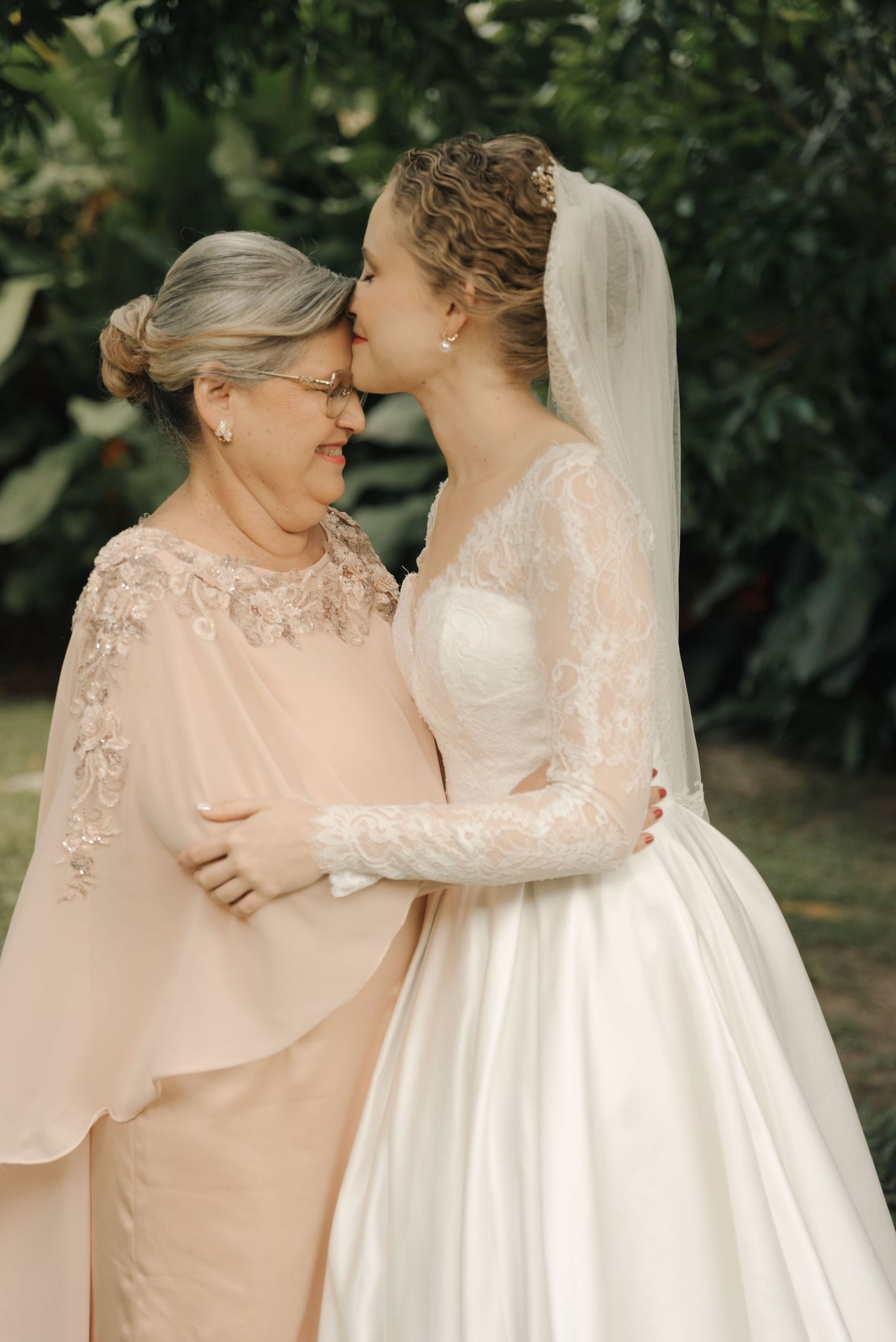 Tender moment between bride in white lace gown and woman in blush dress.