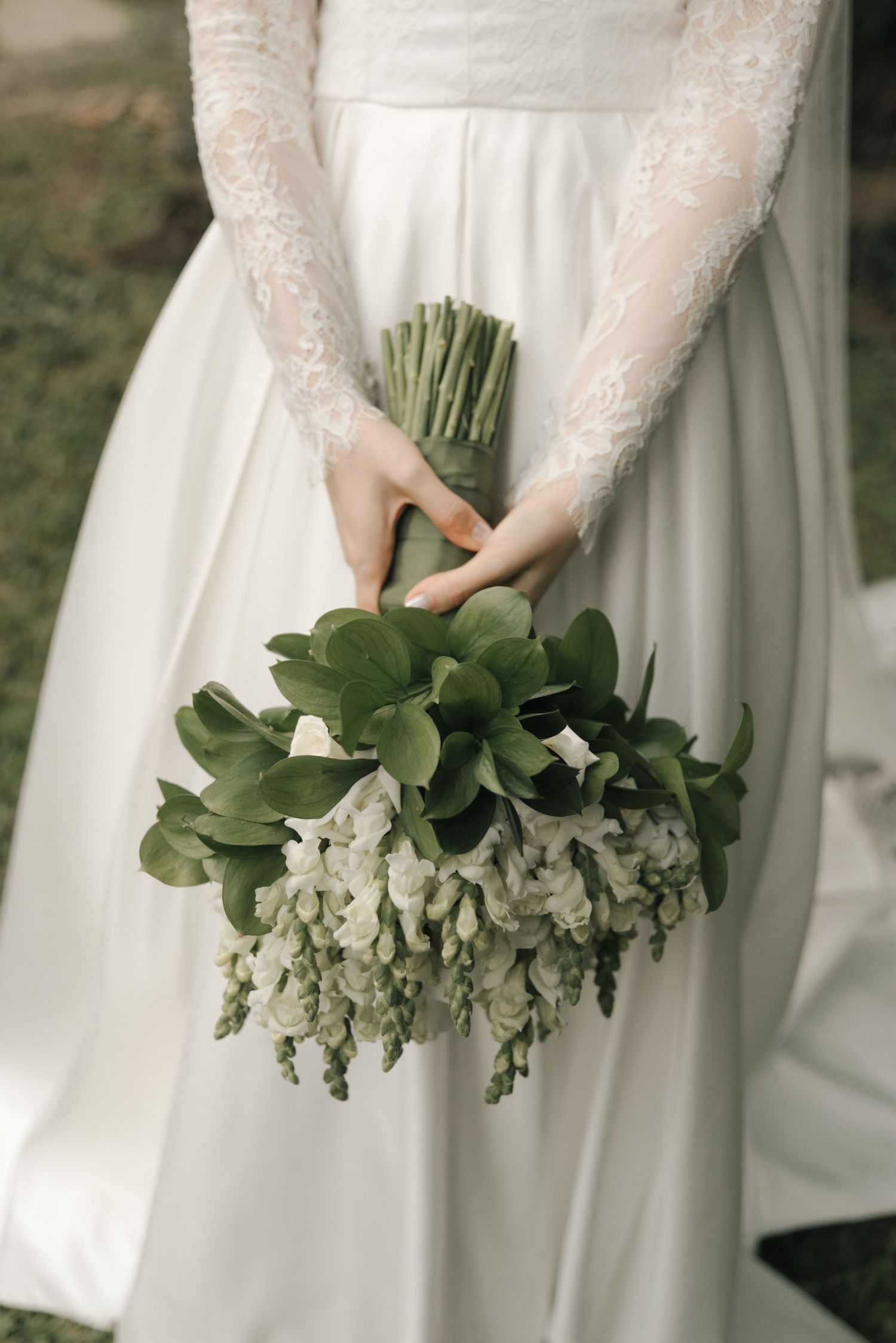 Wedding bouquet with white flowers and green foliage held against a lace-sleeved white dress.