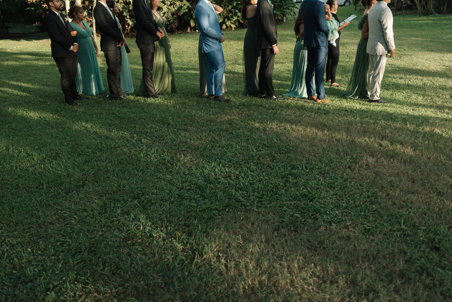 Wedding party members in teal outfits standing in a row on grass under trees.