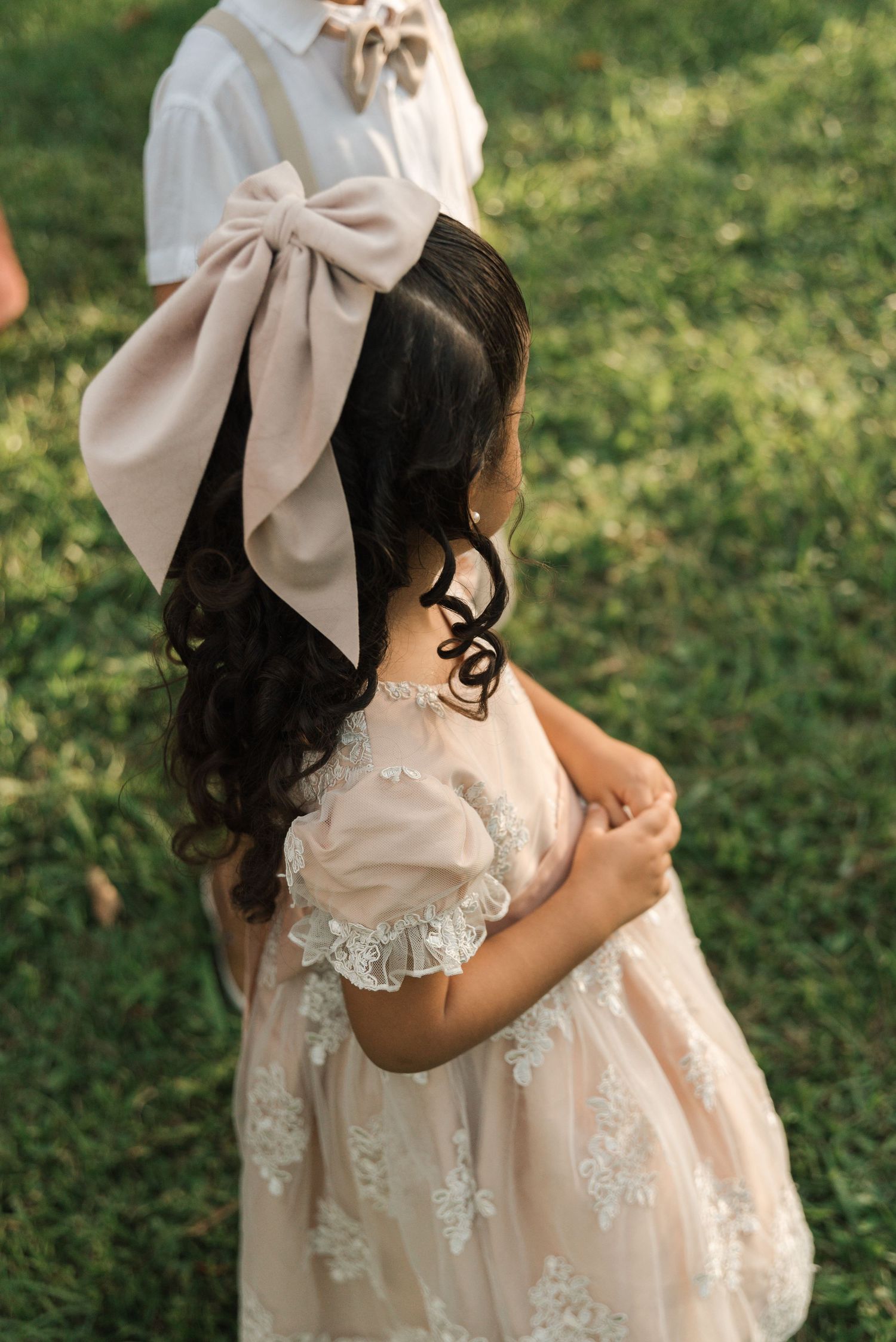 Young flower girl in cream lace dress with large bow hair accessory on grass.