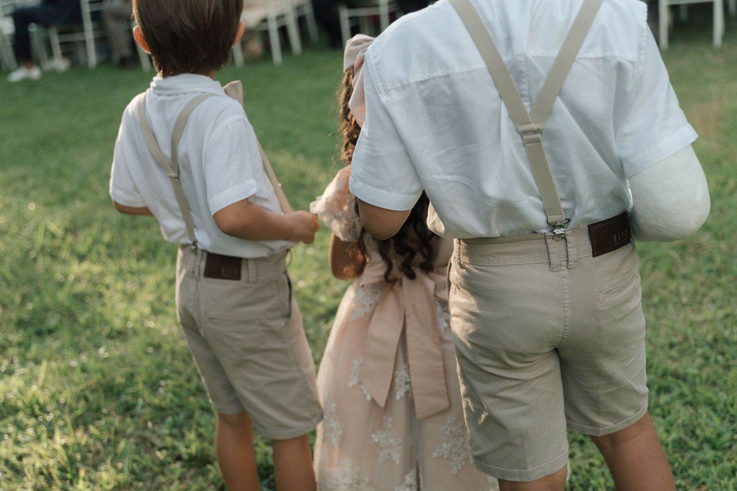 Children in matching khaki shorts and suspenders line up at an outdoor wedding.