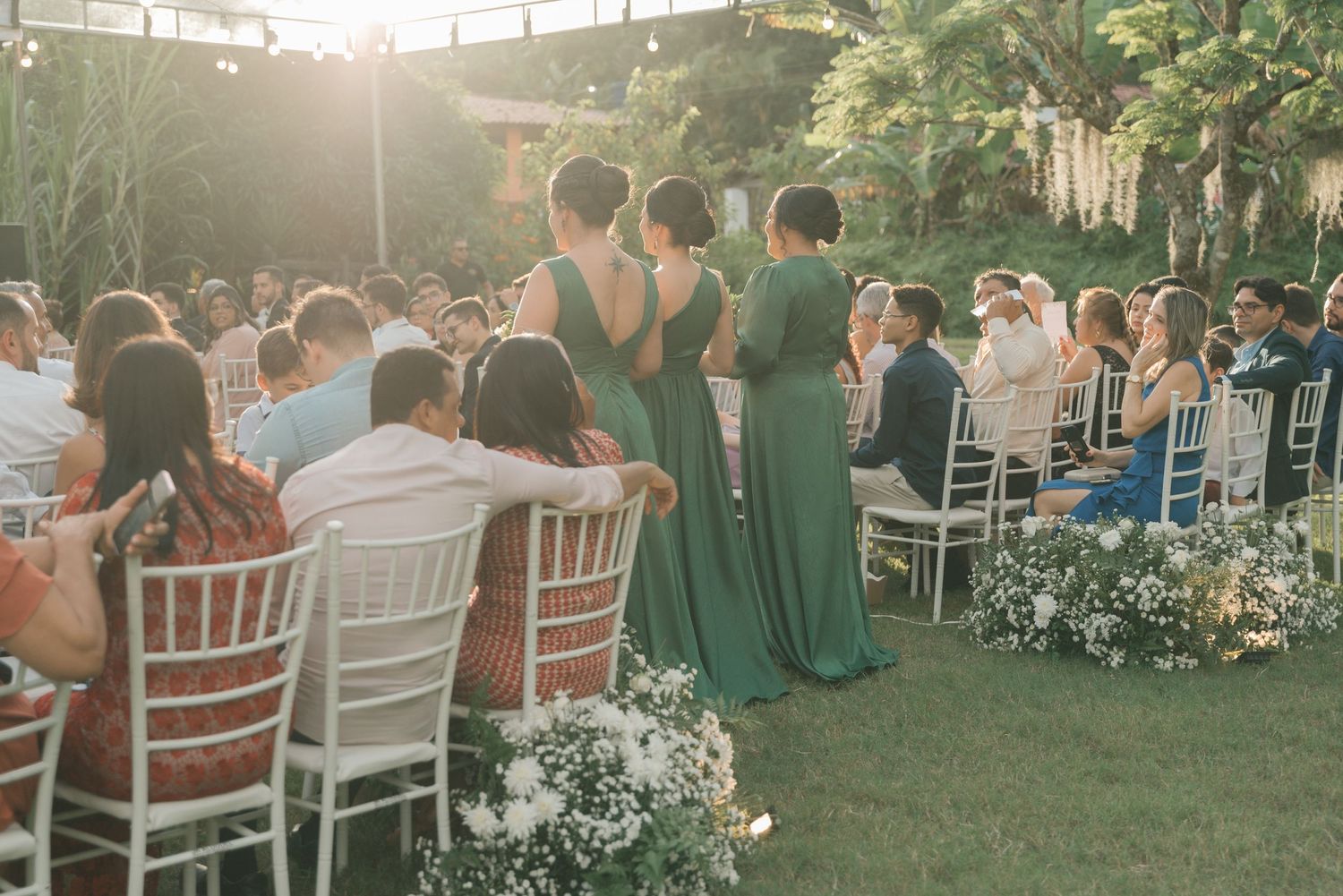 Outdoor wedding ceremony setup with white chairs and green-dressed bridesmaids in sunlit garden.
