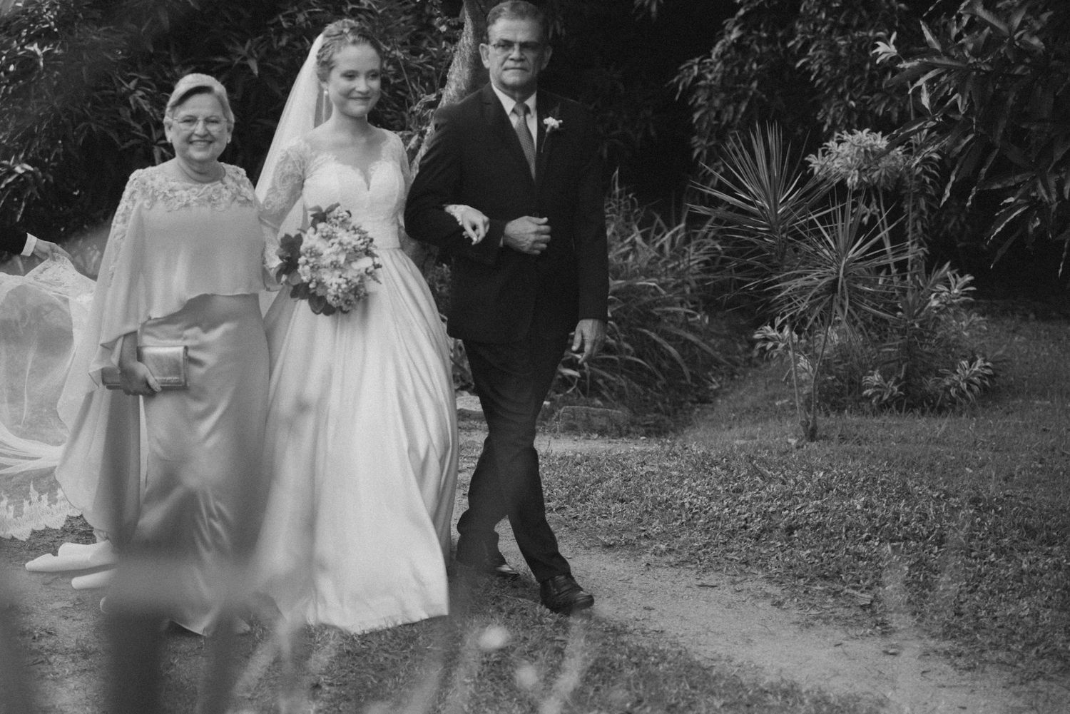 Vintage black and white photo of a bride walking with attendants in a garden setting.