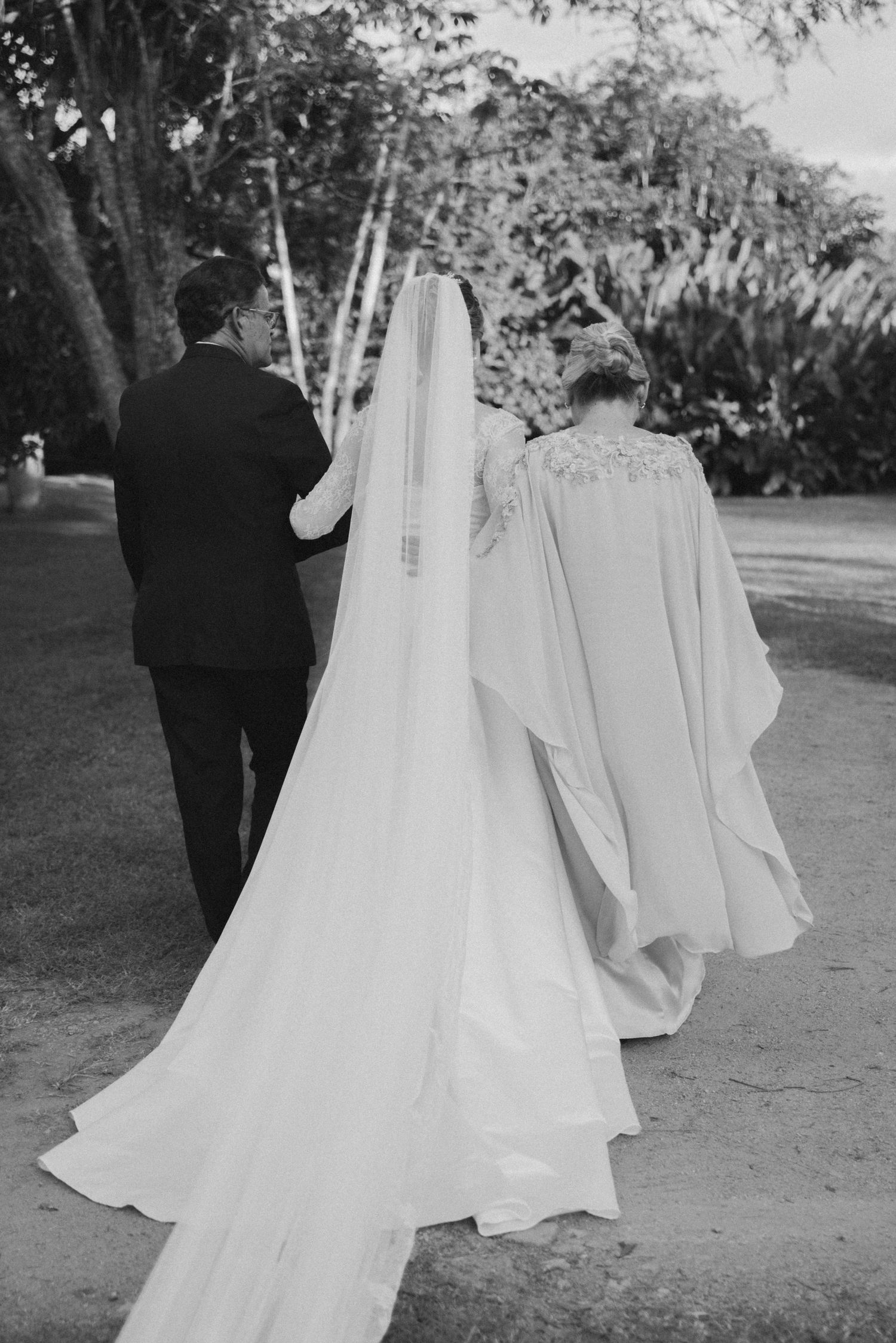 Black and white photo of a bride in flowing veil walking with attendants on garden path.