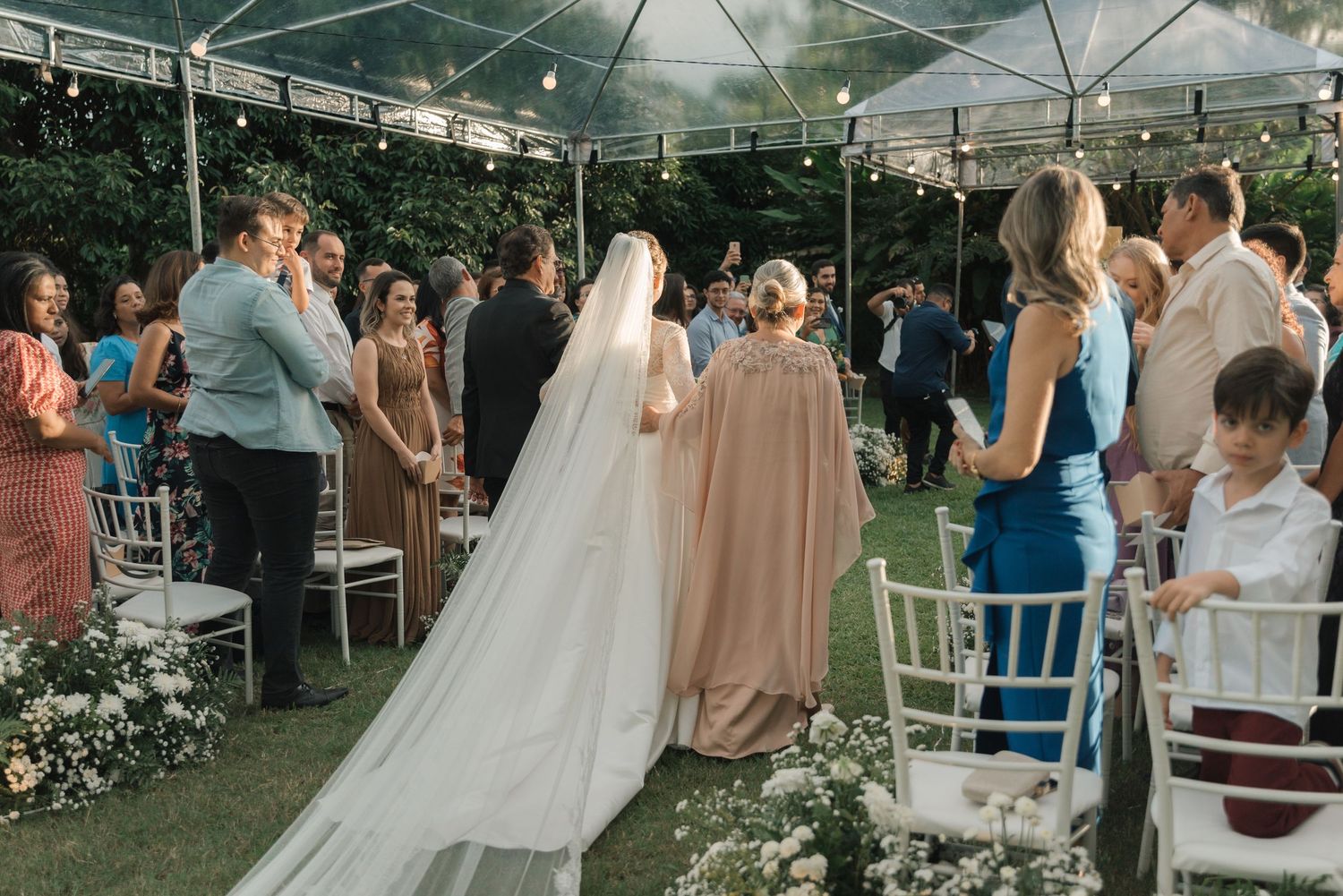 Outdoor wedding ceremony with guests seated on white chairs under clear tent.