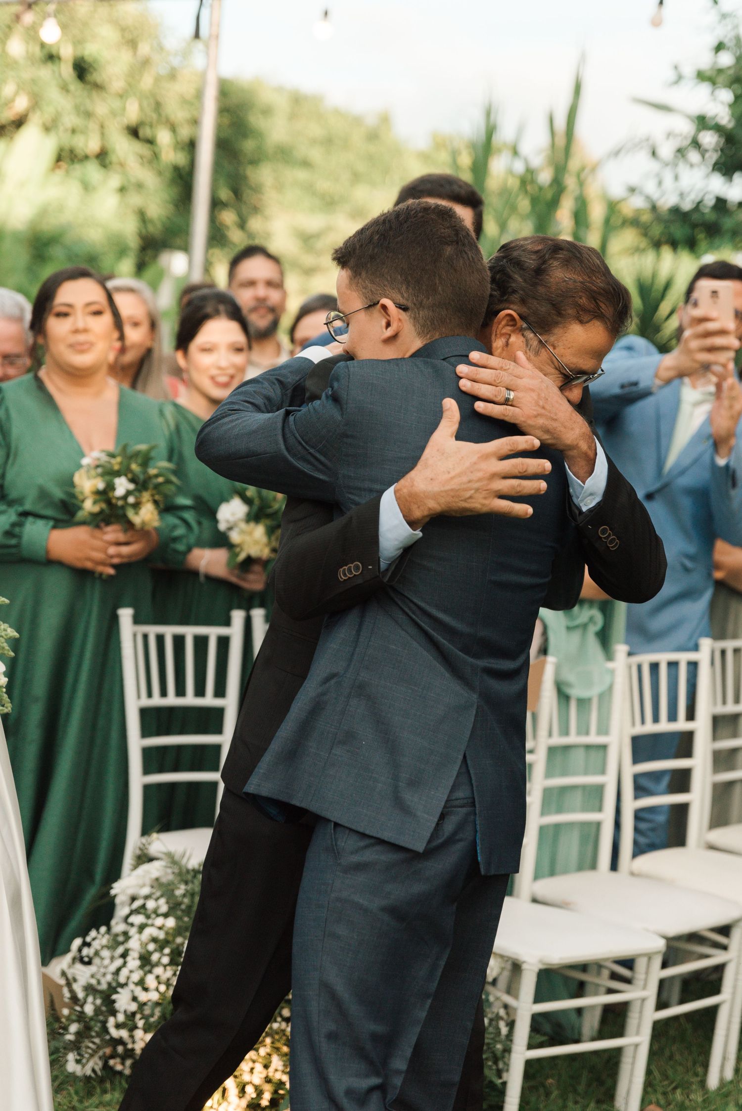 Emotional embrace at outdoor wedding ceremony with guests in formal attire watching.