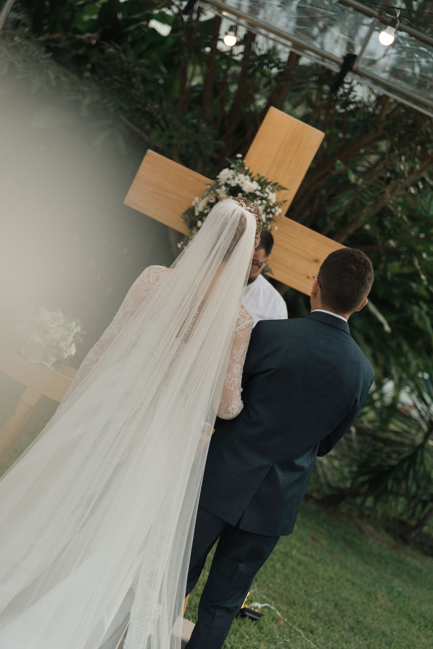 Wedding couple standing before a wooden cross adorned with flowers during outdoor ceremony.