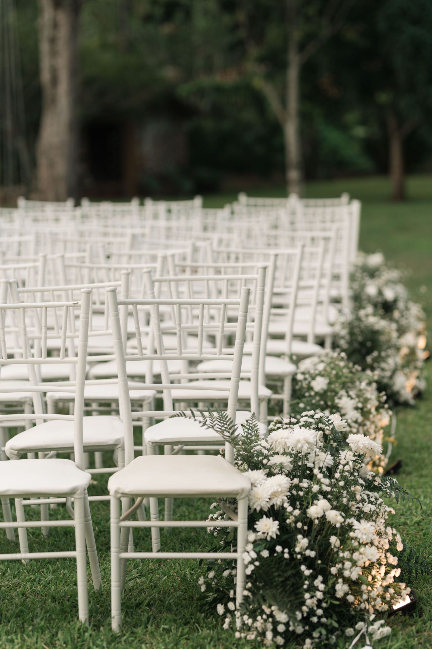 White garden chairs lined up with delicate floral arrangements for outdoor wedding ceremony.
