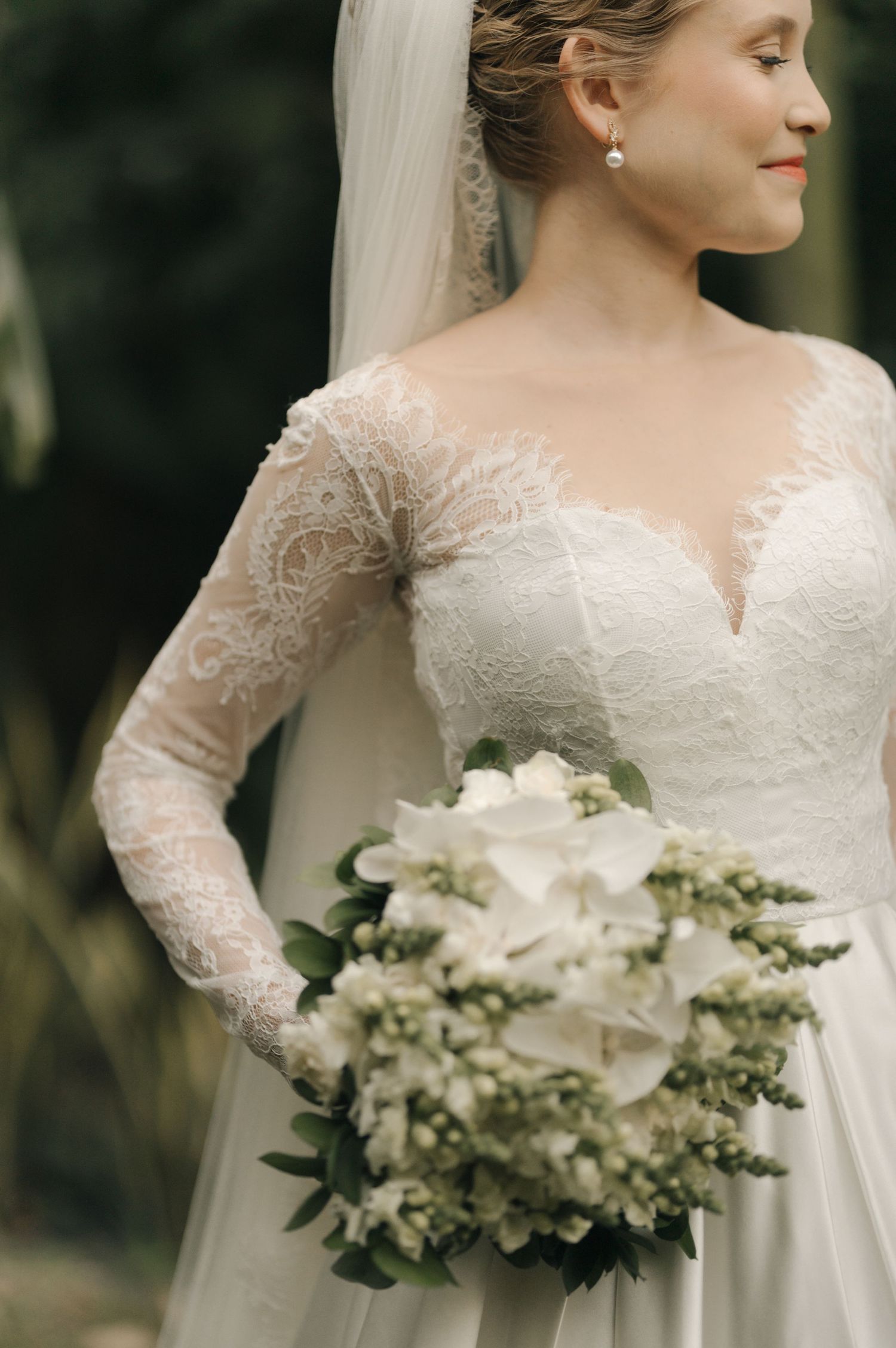 Bride in long-sleeve lace wedding dress holding white bouquet in garden setting.