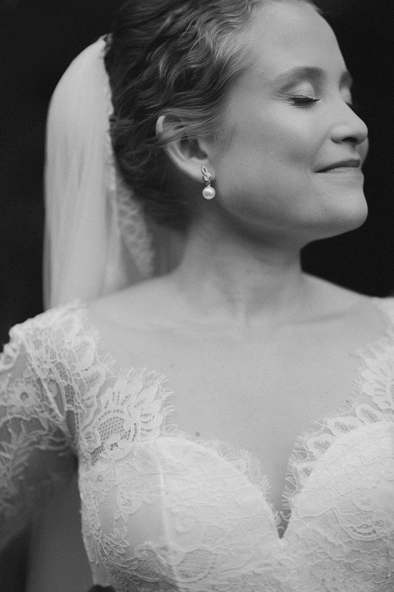 A black and white close-up portrait of a bride wearing a lace wedding dress and pearl earrings with her eyes closed.