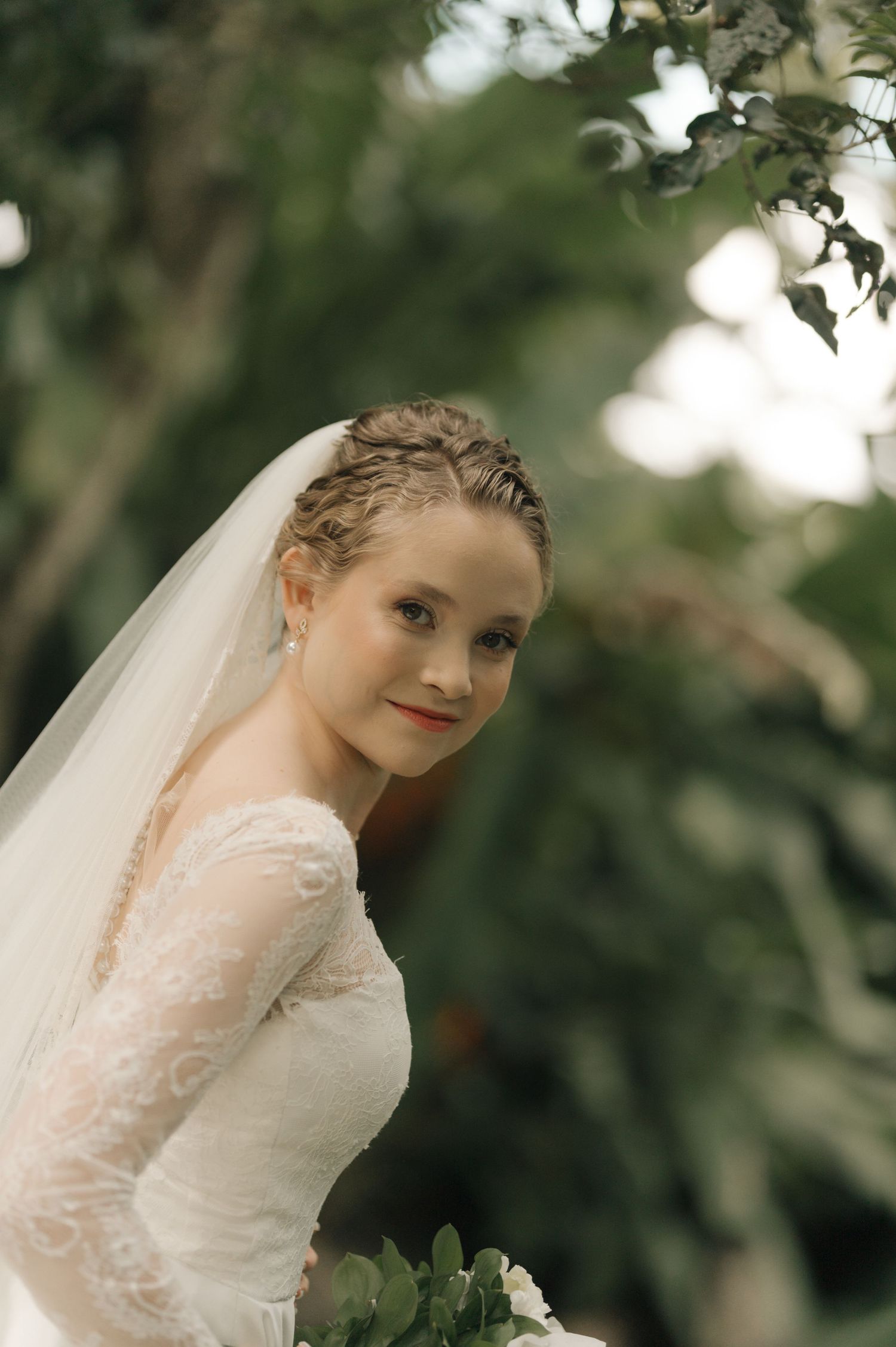 A bride in a long-sleeved lace wedding dress poses outdoors against a natural garden backdrop.