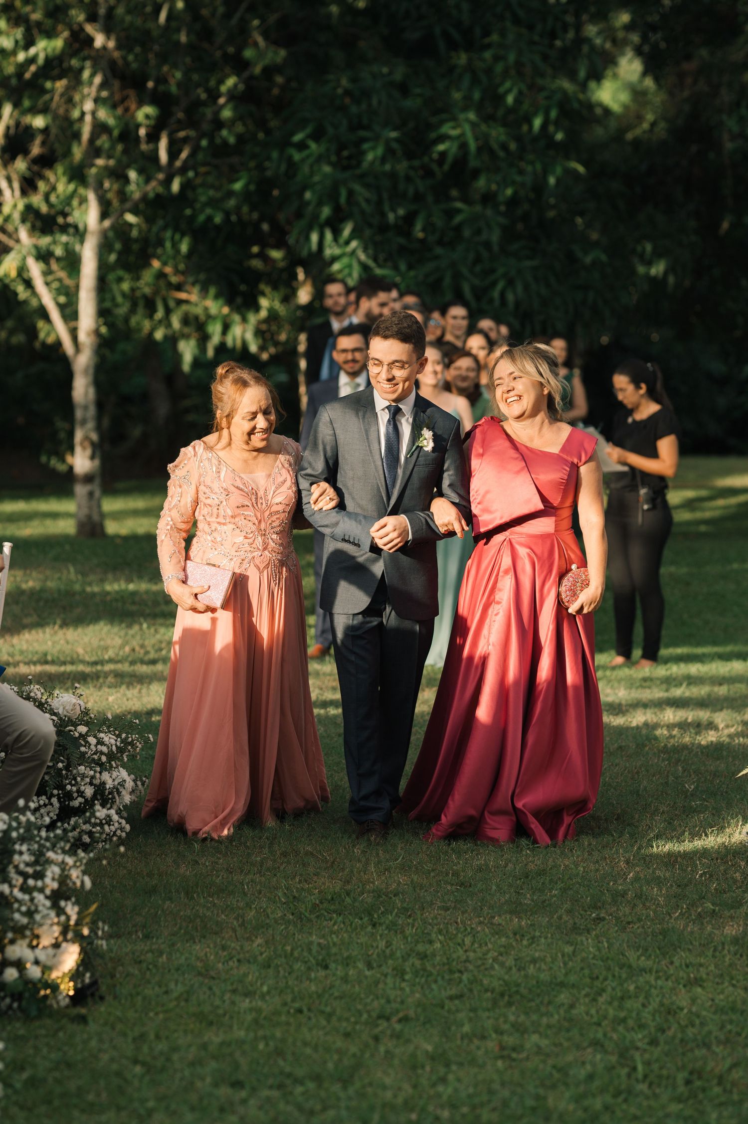 Three wedding guests in coral and pink formal dresses walk arm in arm through a garden setting.