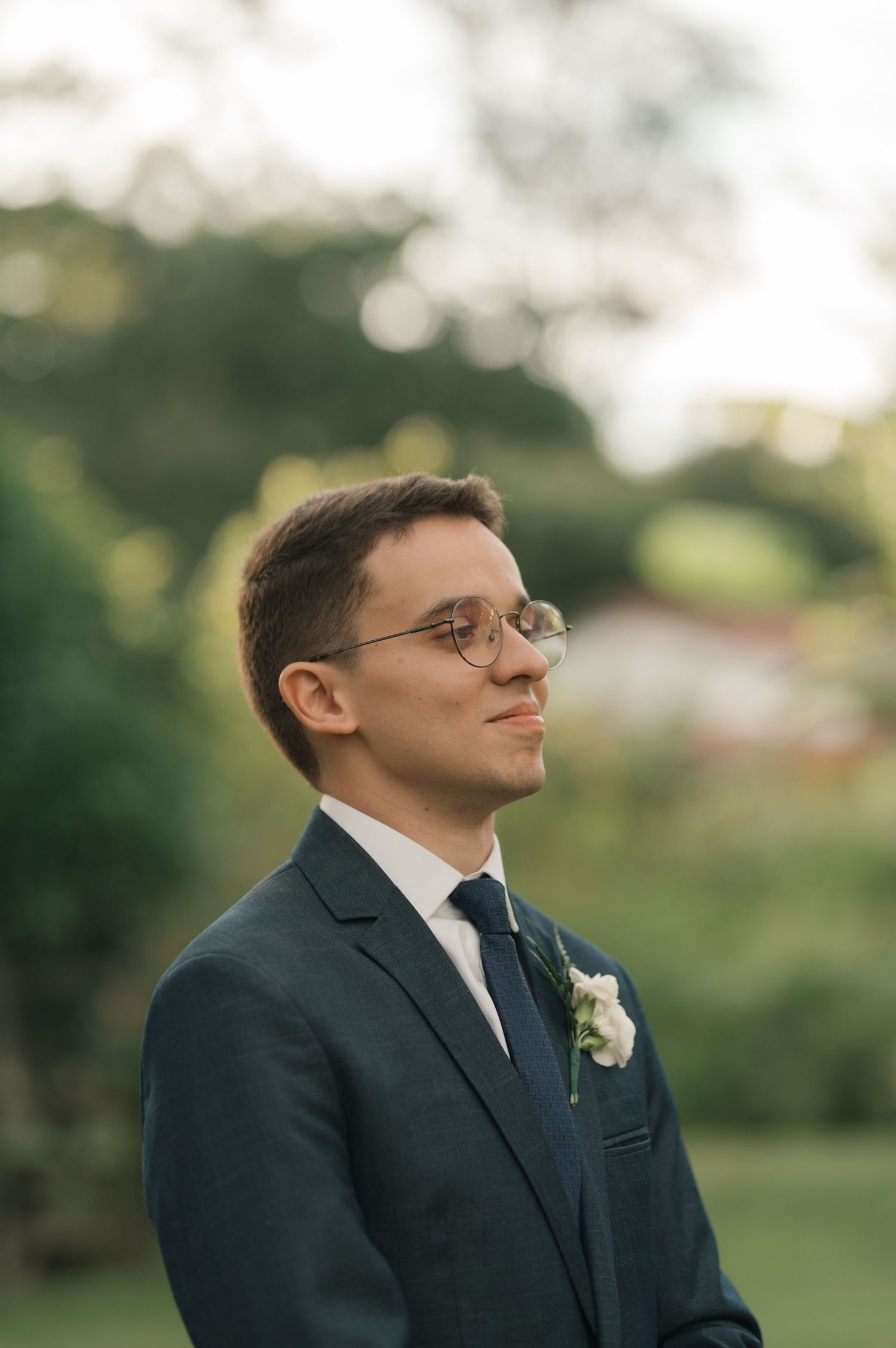 A groom in a navy suit with glasses and boutonniere stands in an outdoor garden setting.