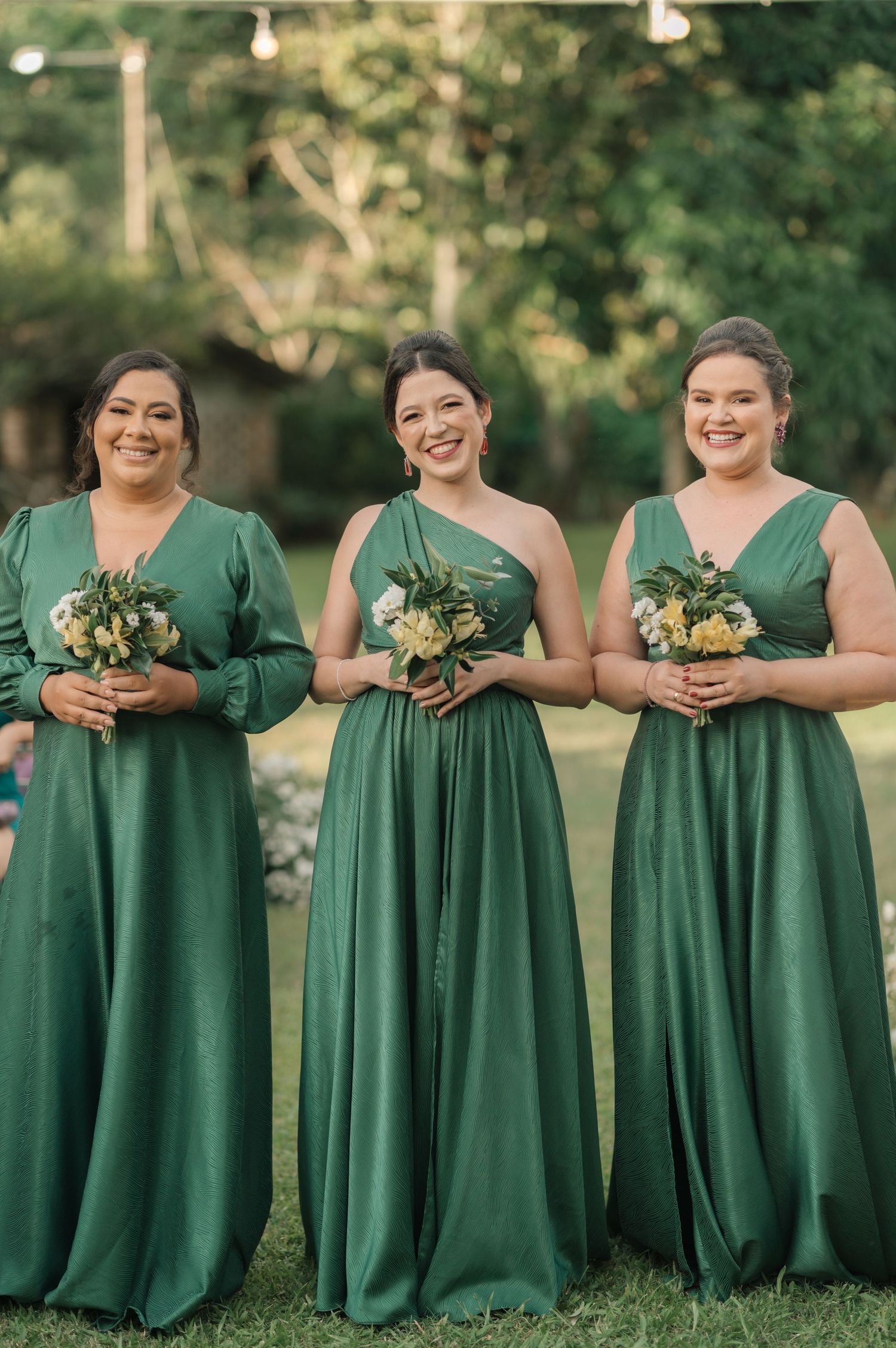 Three bridesmaids in matching emerald green dresses hold bouquets while standing outdoors.