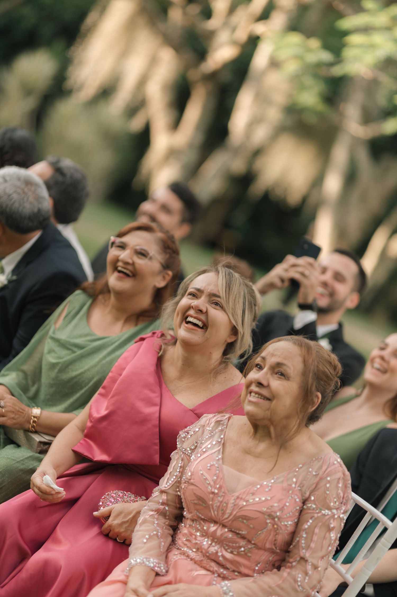 Wedding guests in formal attire laugh joyfully while seated at an outdoor ceremony.