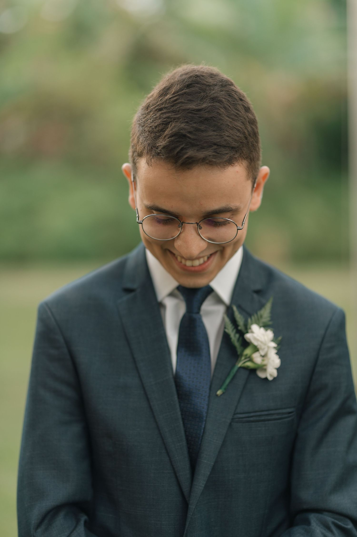 A groom in a navy suit and glasses smiles warmly while wearing a white floral boutonniere.