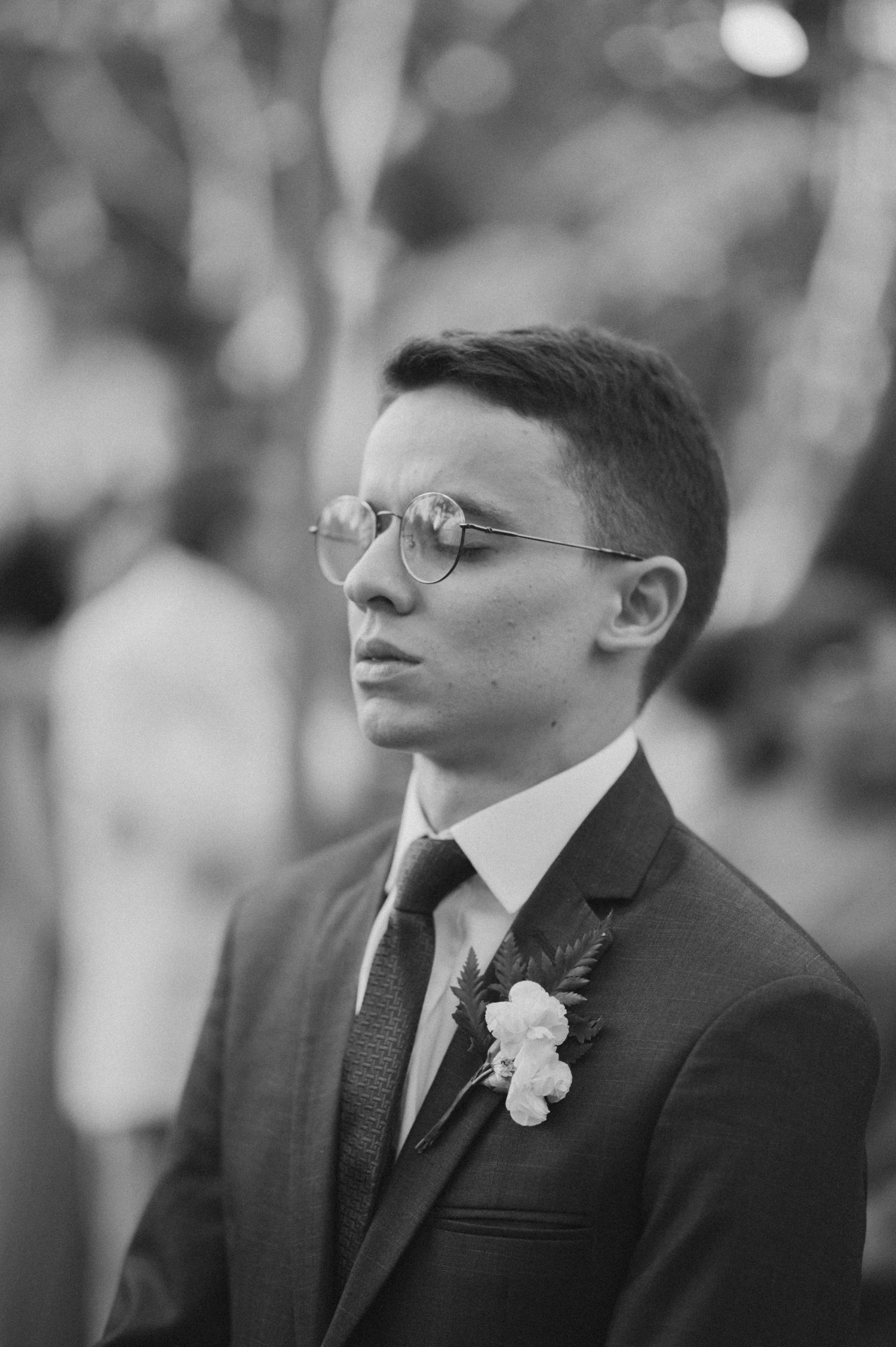 A black and white portrait of a groom in a suit and glasses with a floral boutonniere.