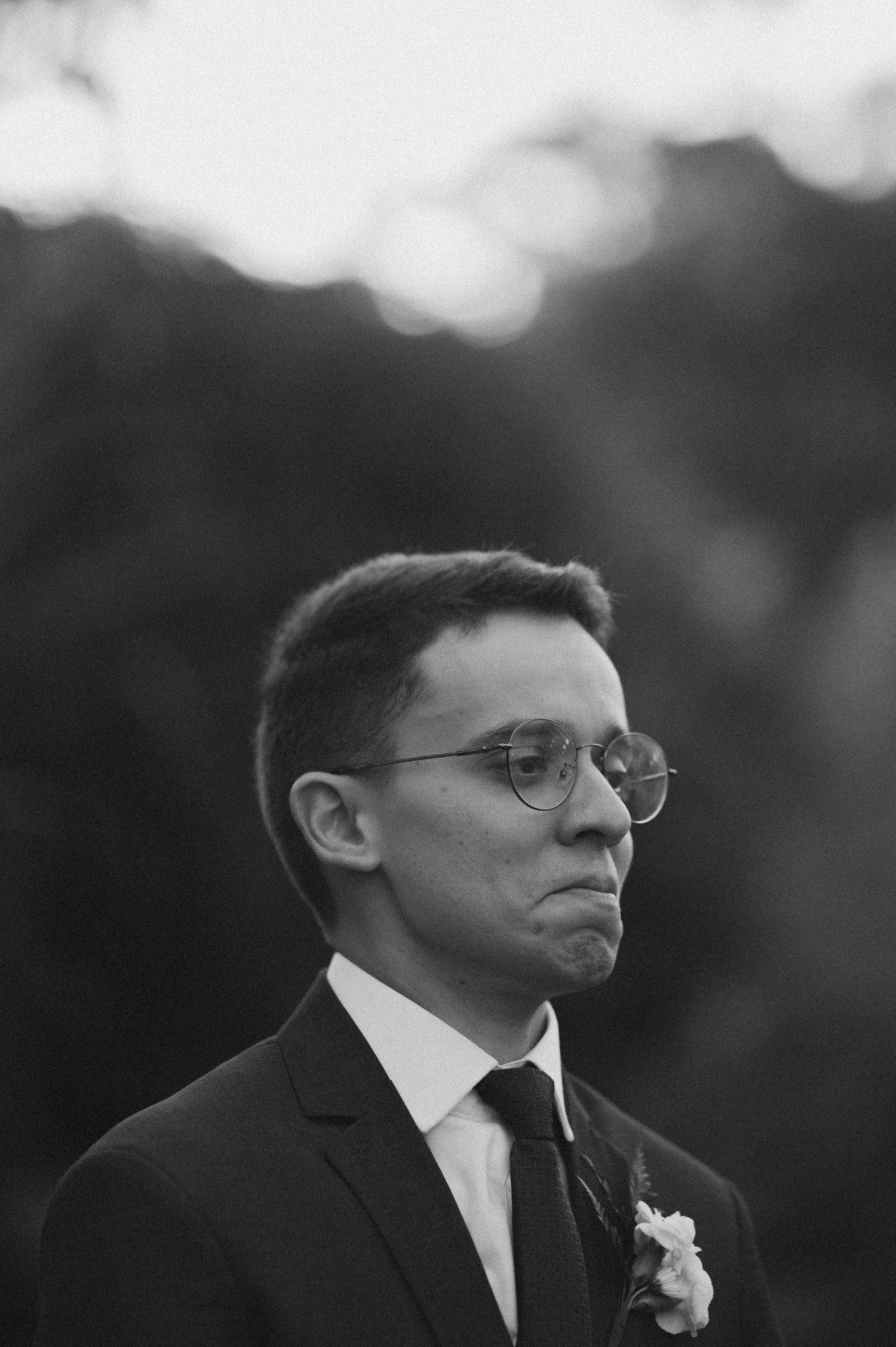 Black and white portrait of a groom wearing glasses and suit with a boutonniere at an outdoor wedding ceremony.