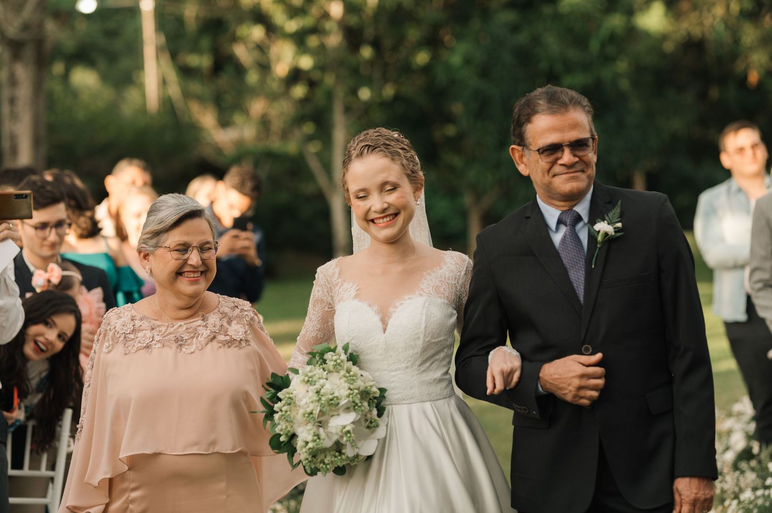 Emotional moment as a bride in white dress walks down the aisle with family members at outdoor wedding.