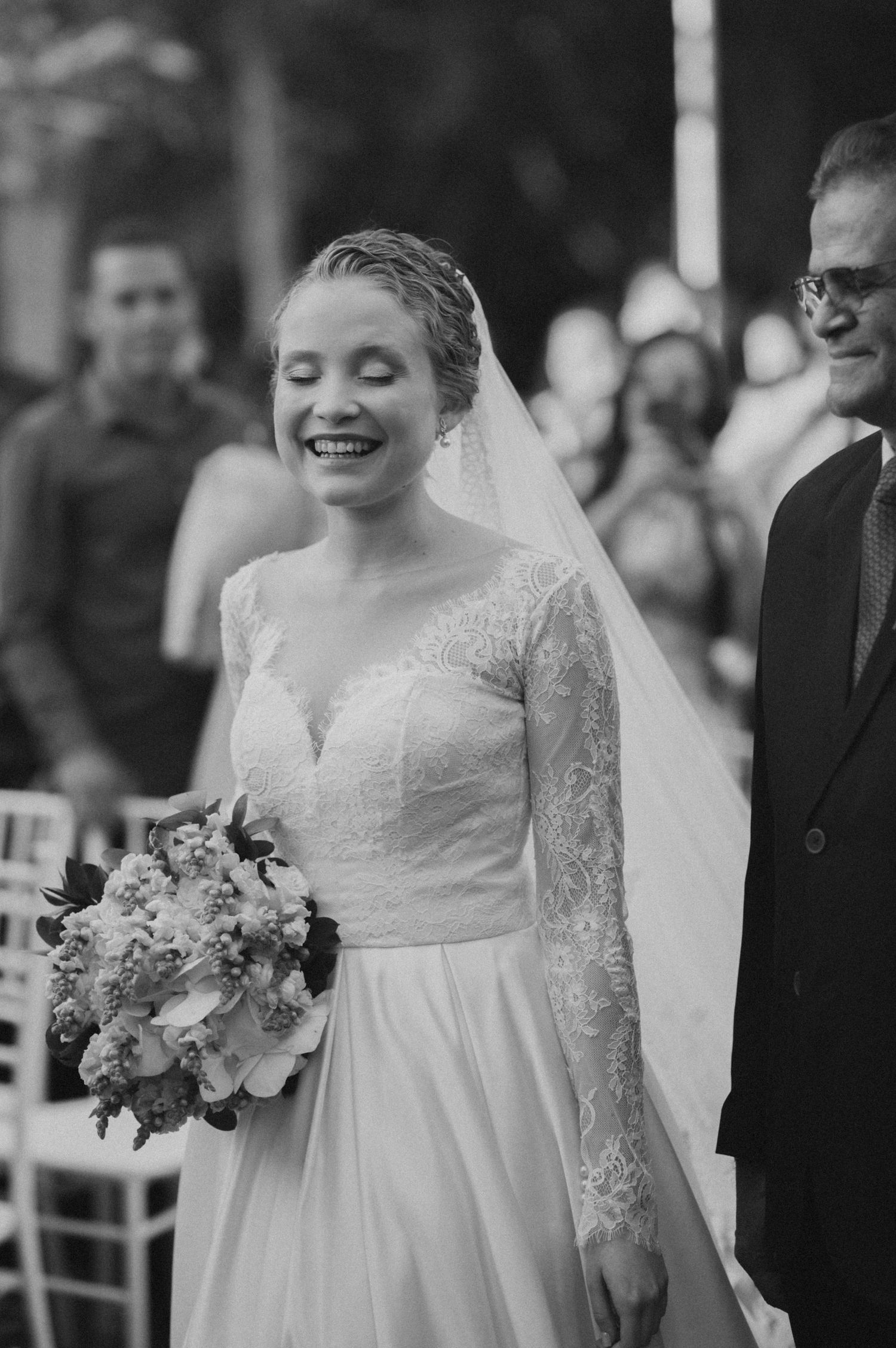 Joyful bride in lace wedding dress holding bouquet during outdoor ceremony.