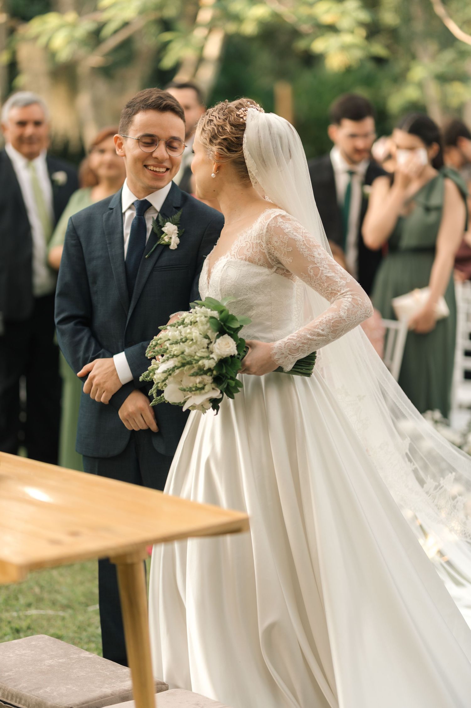 Newlyweds share joyful exchange during outdoor wedding with guests in background.