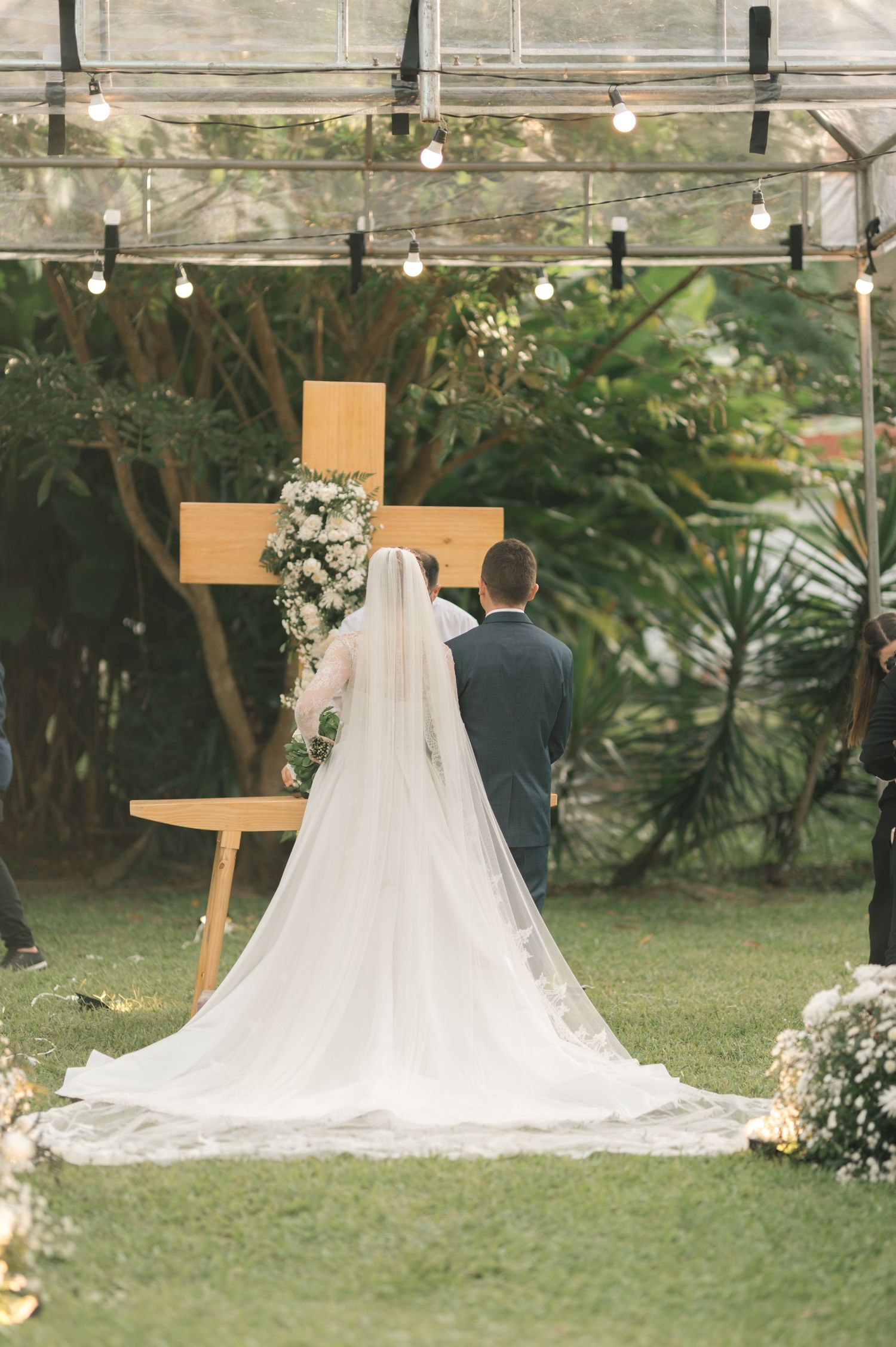 A couple stands before a wooden cross during an outdoor wedding ceremony with string lights and tropical greenery.