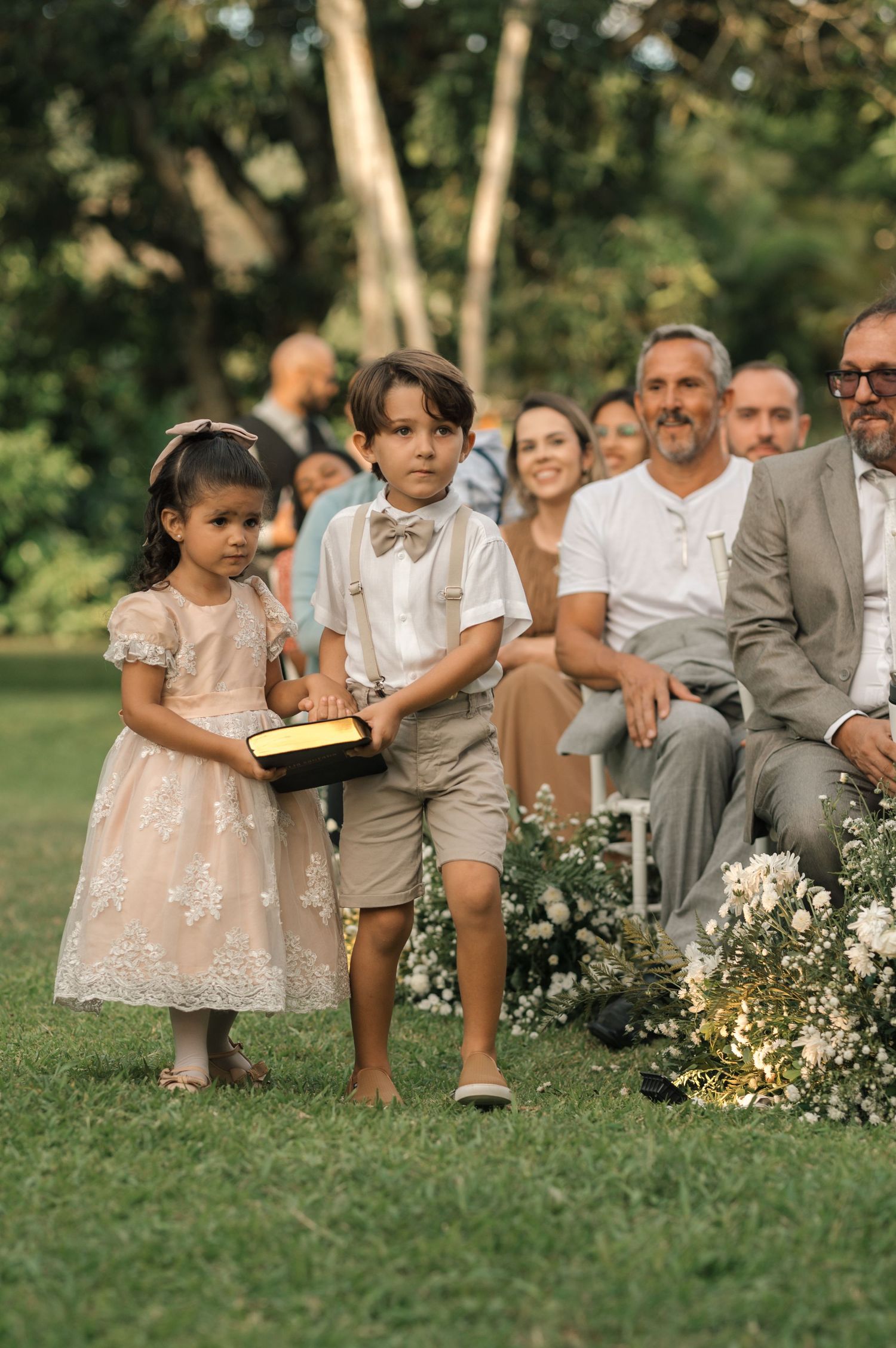 Two young attendants in formal wear stand at the front of a garden wedding ceremony.