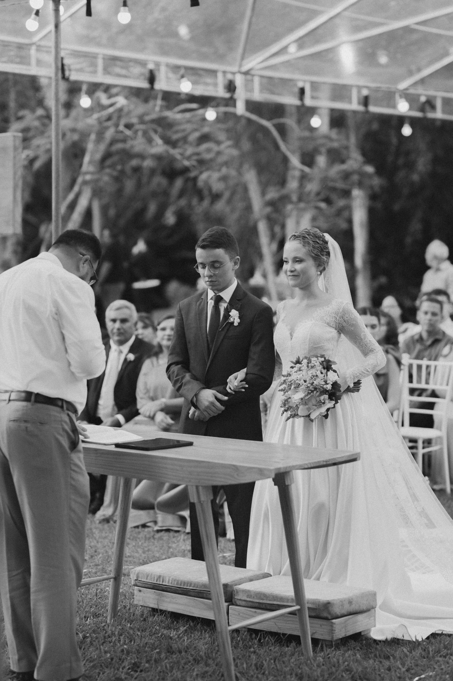 A black and white photo of an outdoor wedding ceremony with string lights and a signing table.