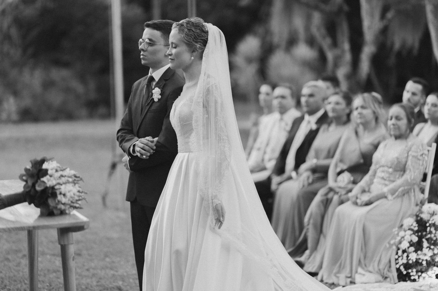 A formal wedding ceremony takes place outdoors with guests seated on white chairs.