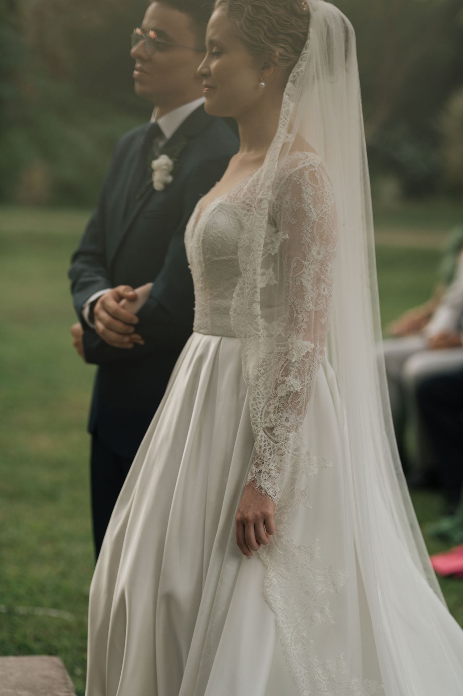 A side view of a bride wearing an elegant lace wedding gown with long sleeves and flowing veil.