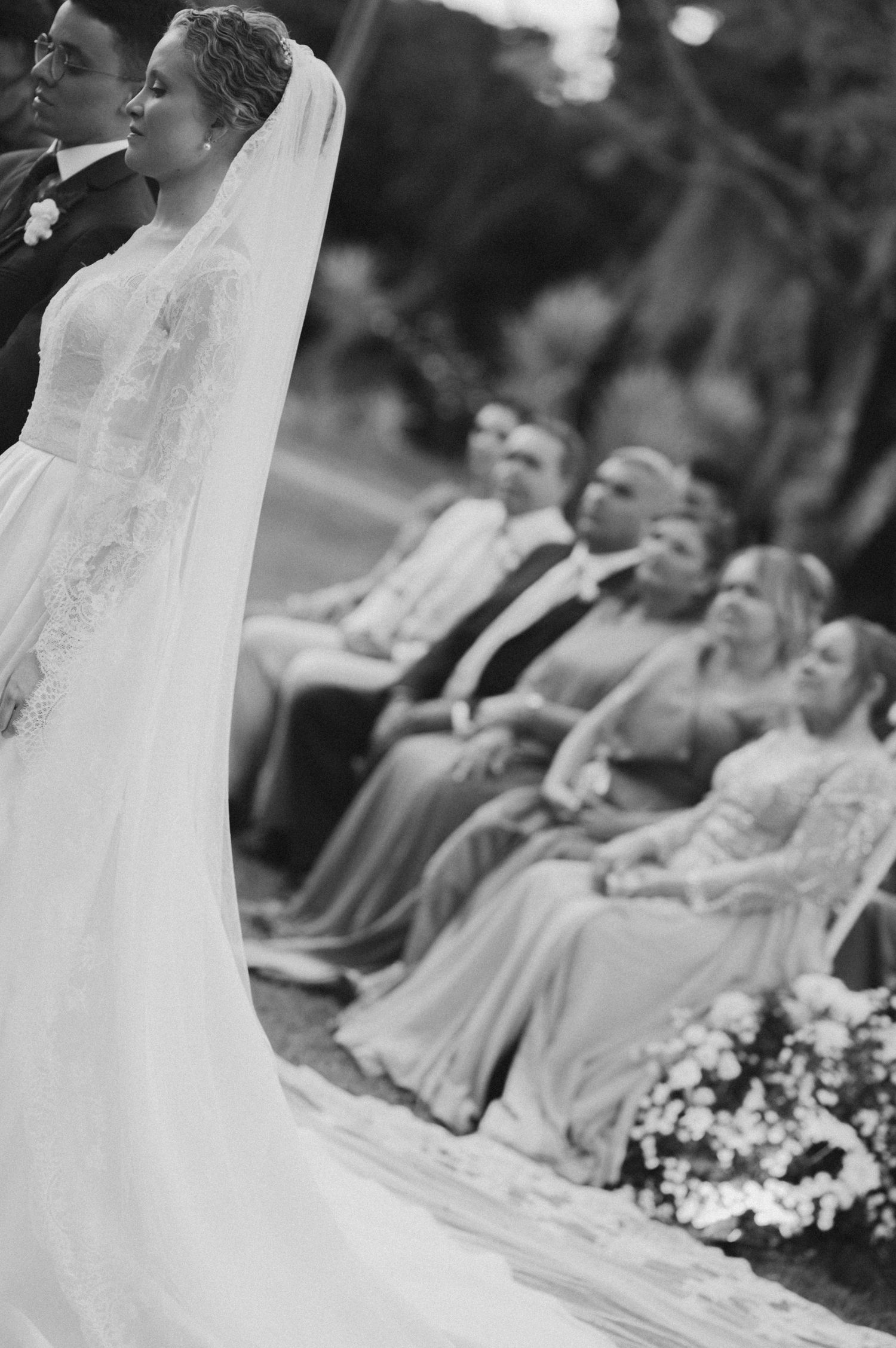 A black and white photo showing a bride in a lace gown with guests seated in rows behind.
