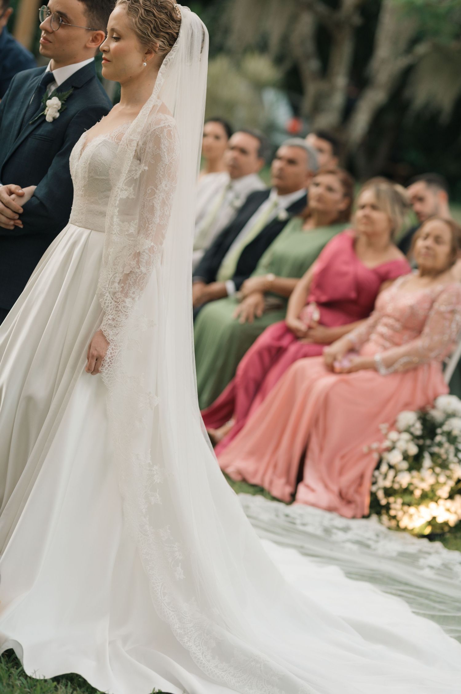 A bride in a classic white gown stands before guests in colorful formal attire at an outdoor ceremony.