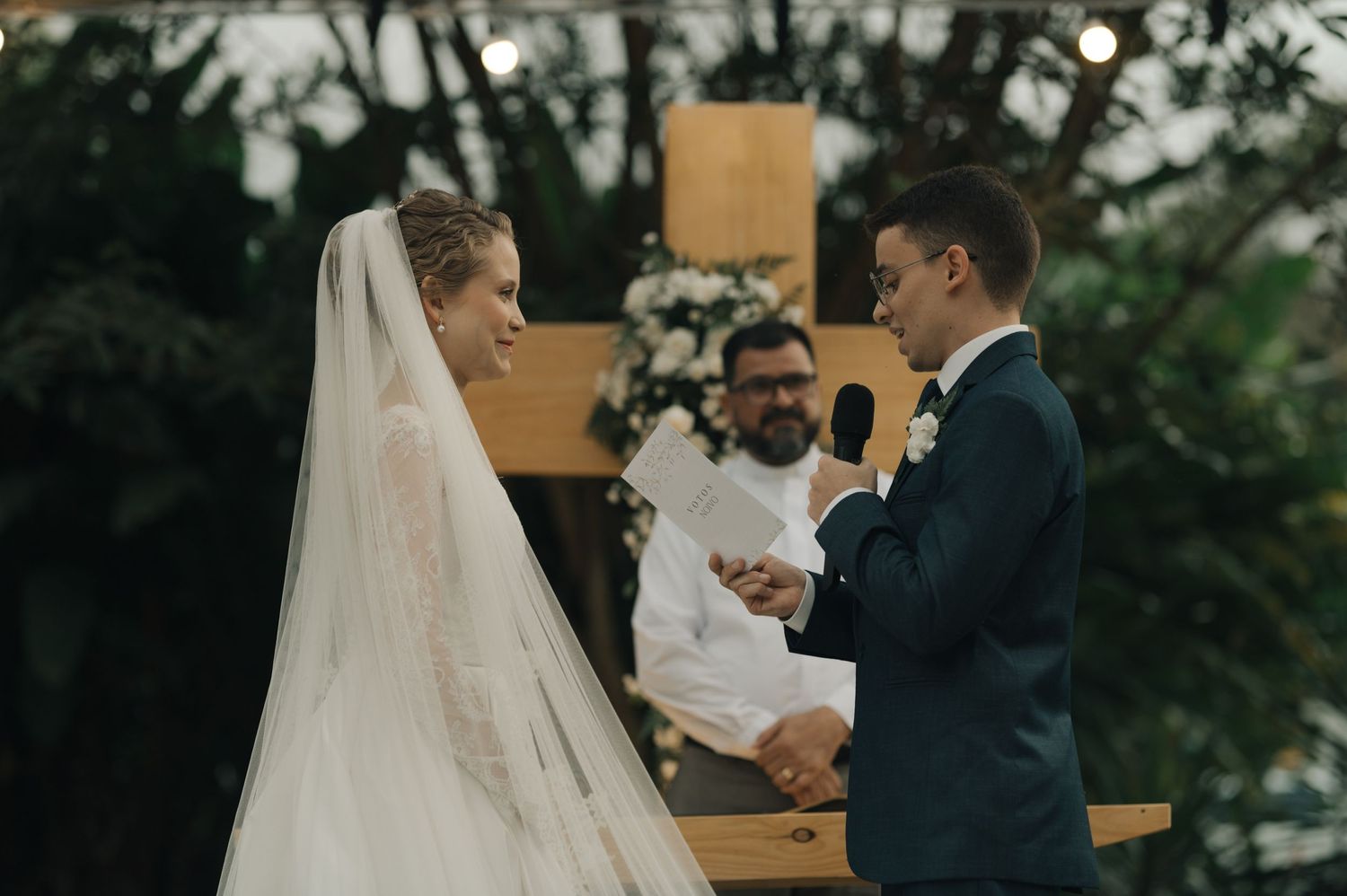 An outdoor wedding ceremony in progress beneath string lights and a wooden cross altar.